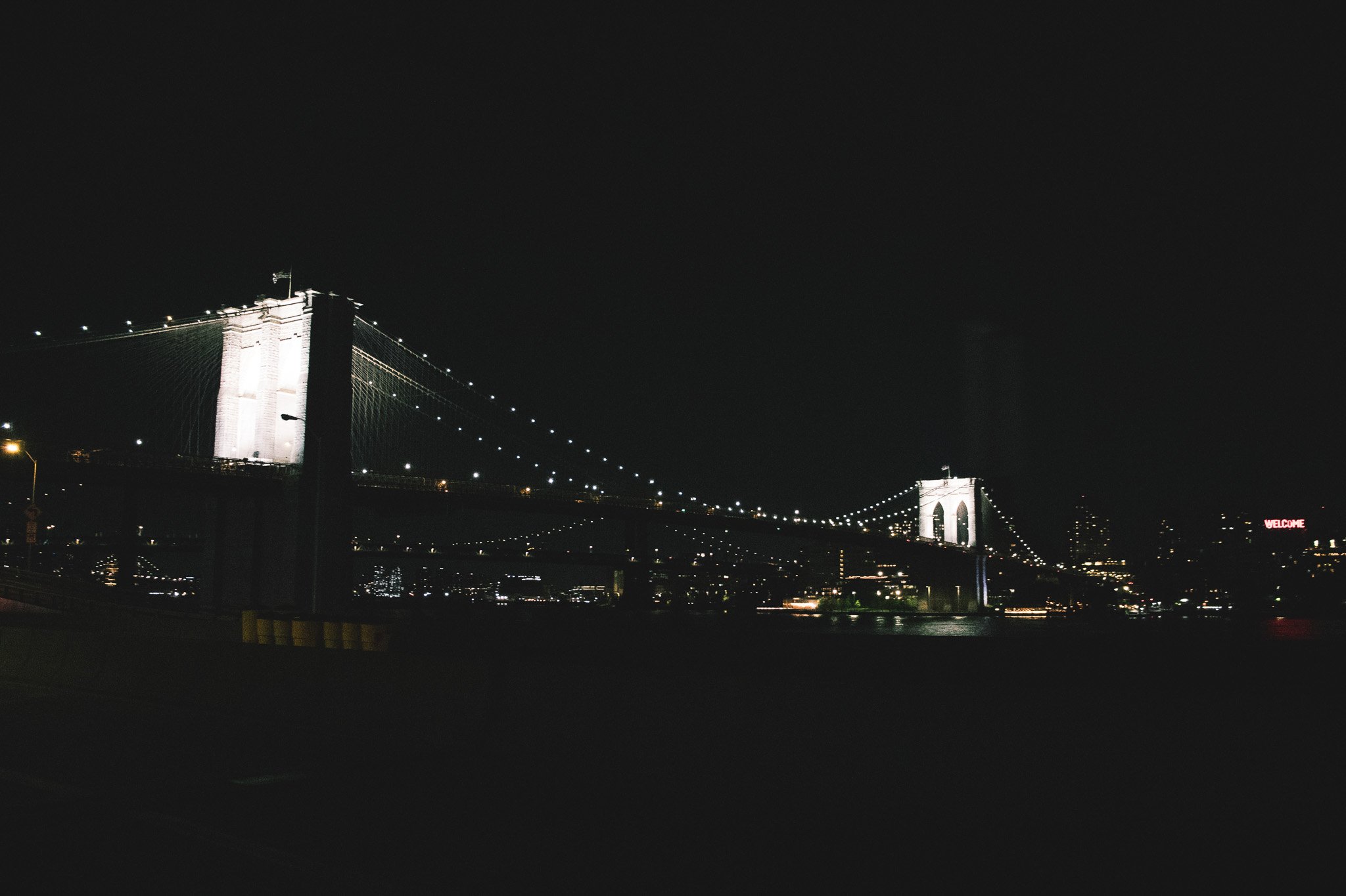 Nighttime view of the Brooklyn Bridge, illuminated with white lights, spanning across a dark river with city lights in the background.
