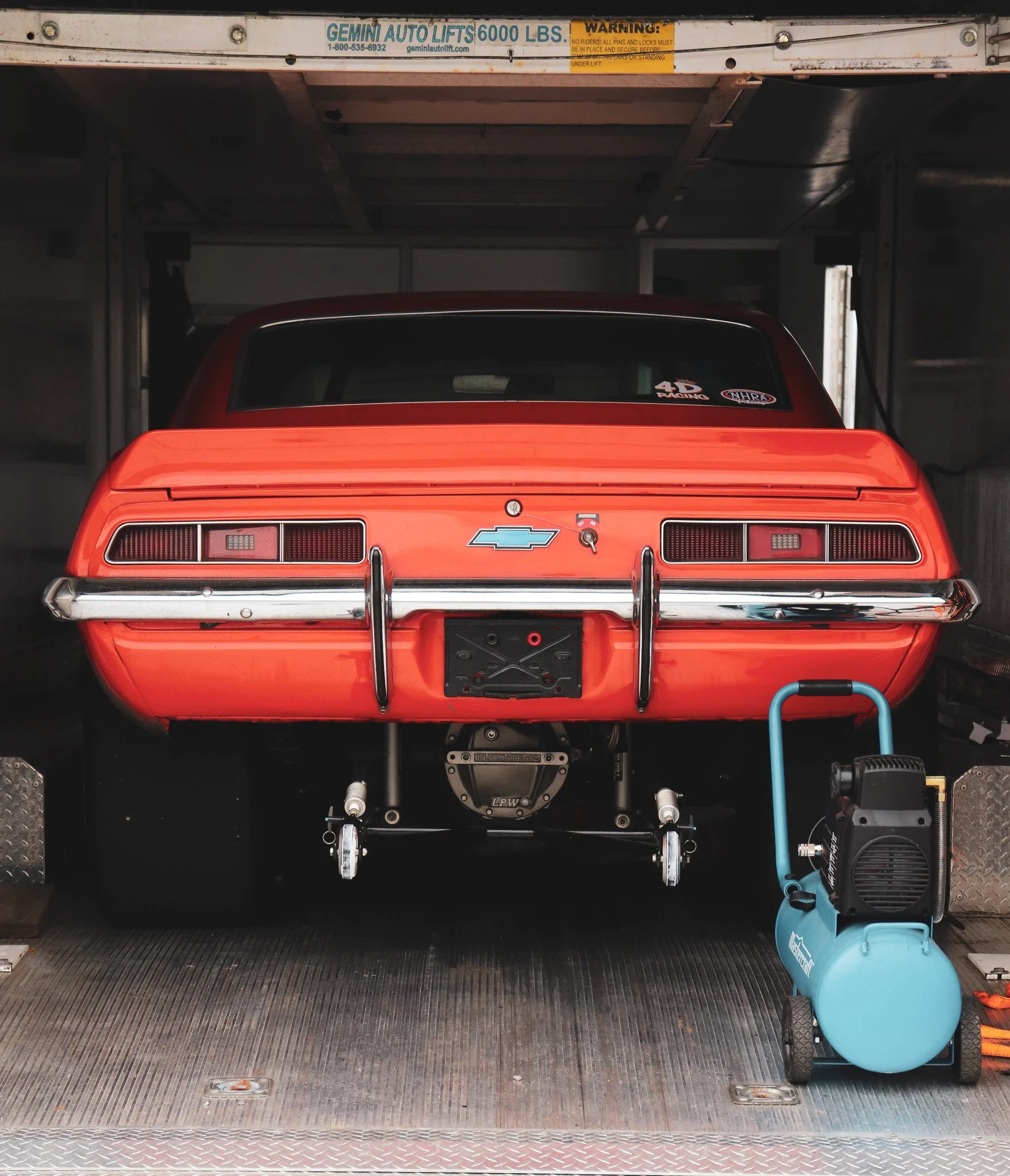 The rear of a bright red vintage Chevrolet Camaro car securely fastened inside a large cargo van or trailer. The vehicle appears to be a drag racing car and is surrounded by equipment, including a blue air compressor on wheels.