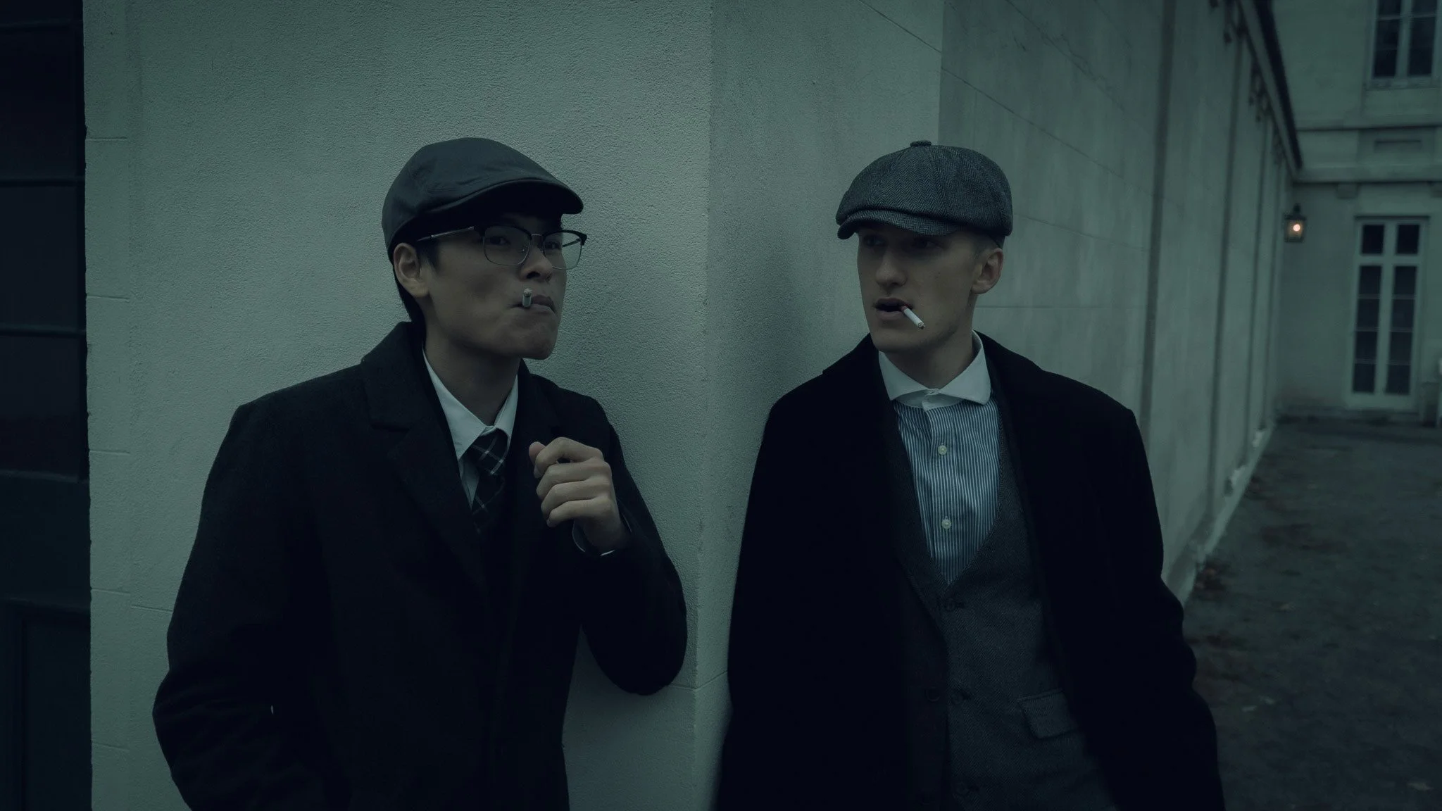 Two men wearing vintage suits and caps, smoking cigarettes and standing against a wall outside a building during dusk.