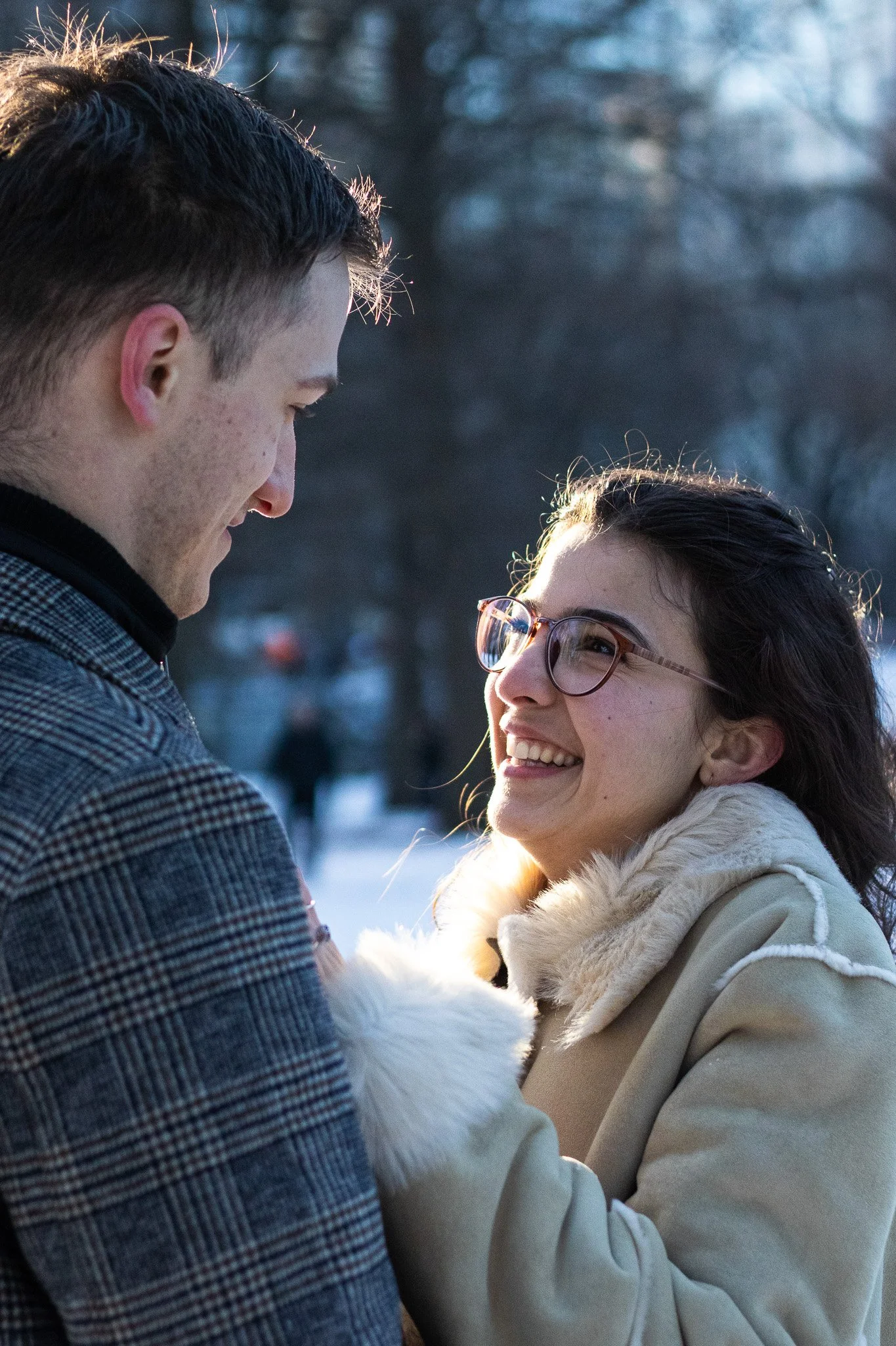 A man and a woman face each other outdoors, smiling and gazing into each other's eyes, with a background of bare trees and soft winter sunlight.