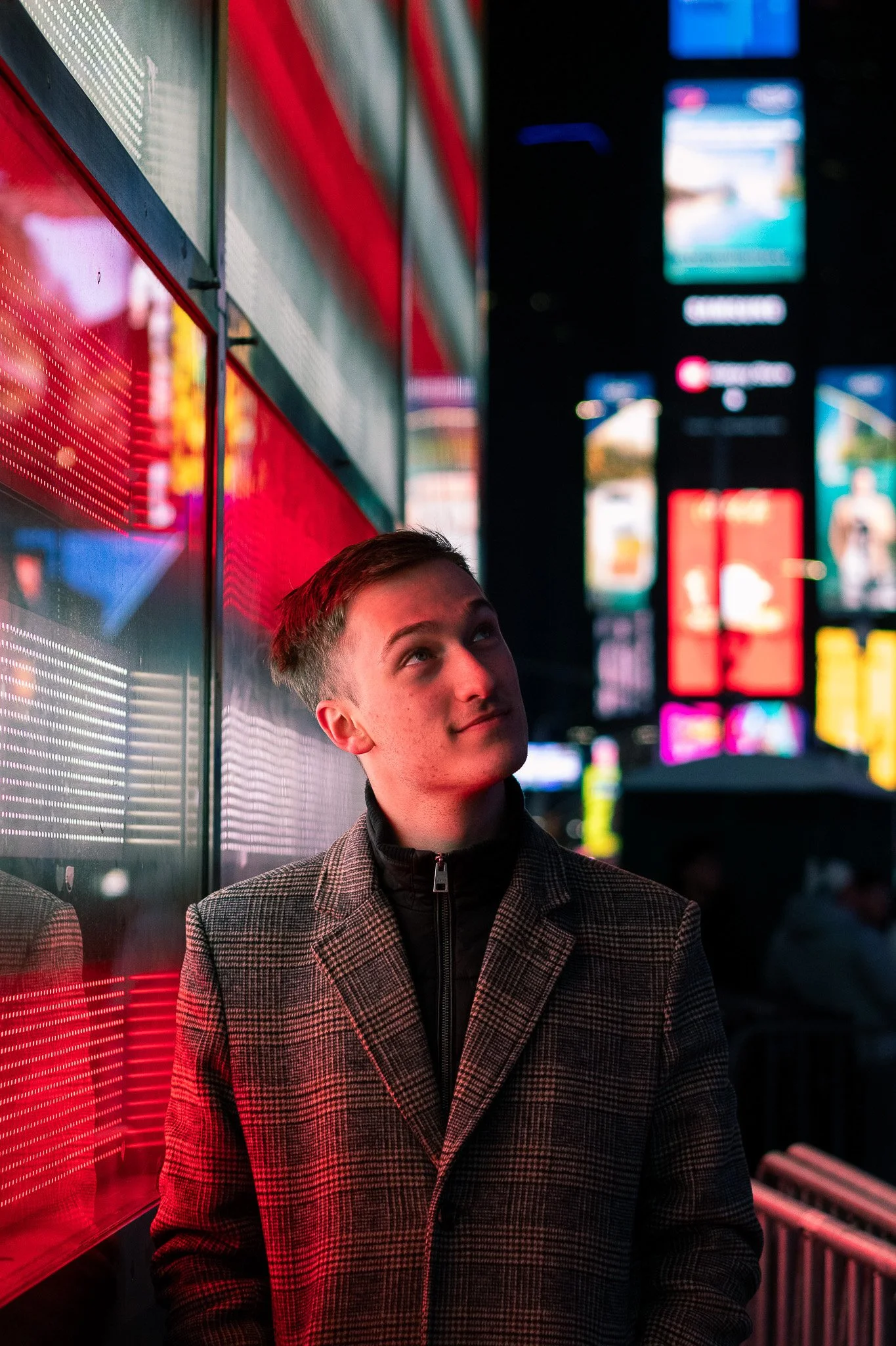 A young man in a checked coat standing at night in a brightly lit urban area with colorful billboards and electronic screens, looking upwards.