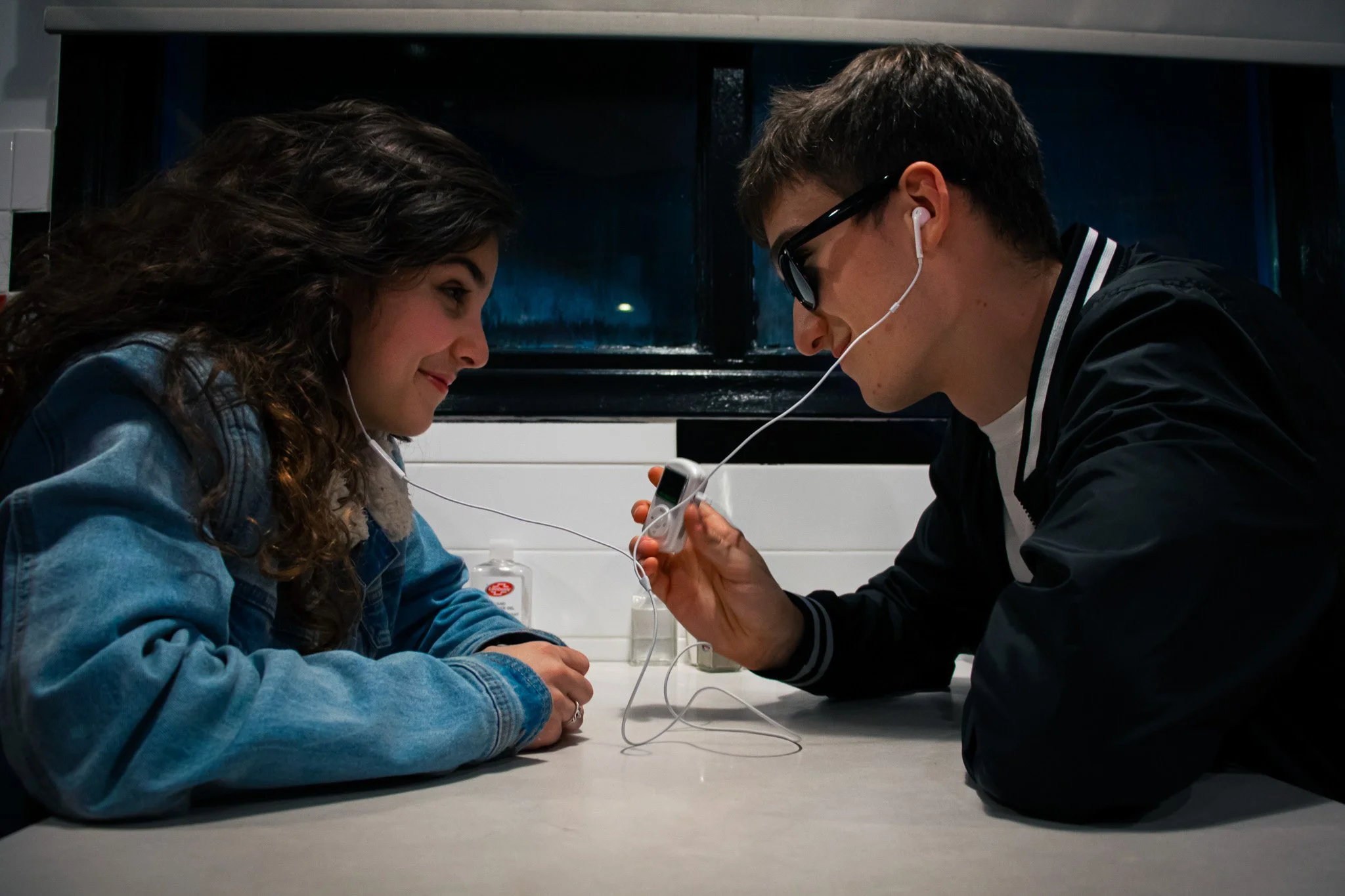 A young woman and a young man face each other across a table, both smiling slightly and leaning toward each other. The woman has long, curly dark hair and is wearing a denim jacket. The man has short dark hair, is wearing sunglasses, and hearing whit