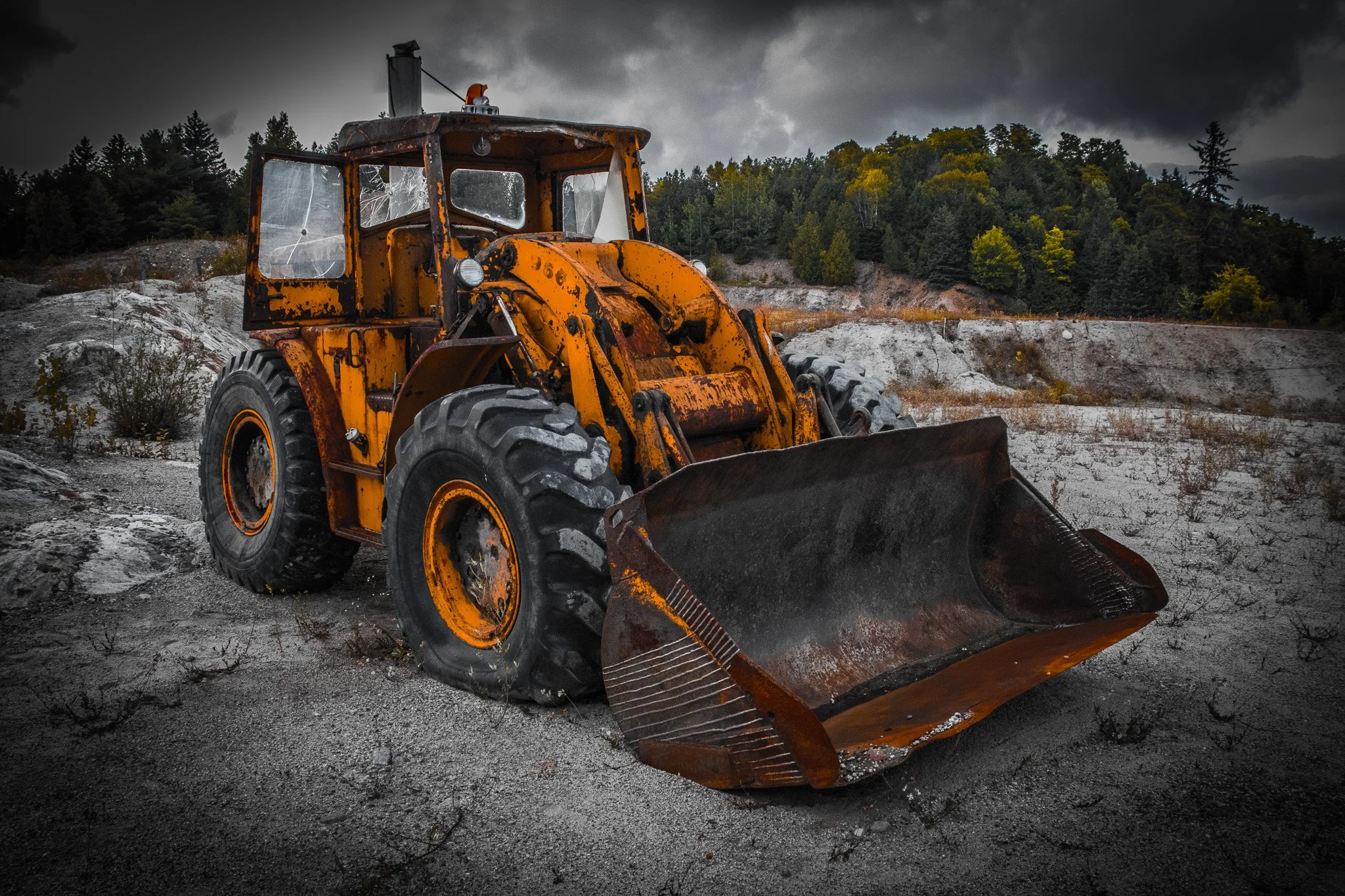 A large yellow bulldozer with shattered windows parked on a rocky, barren landscape under a dark, cloudy sky, with a forested hillside in the background.