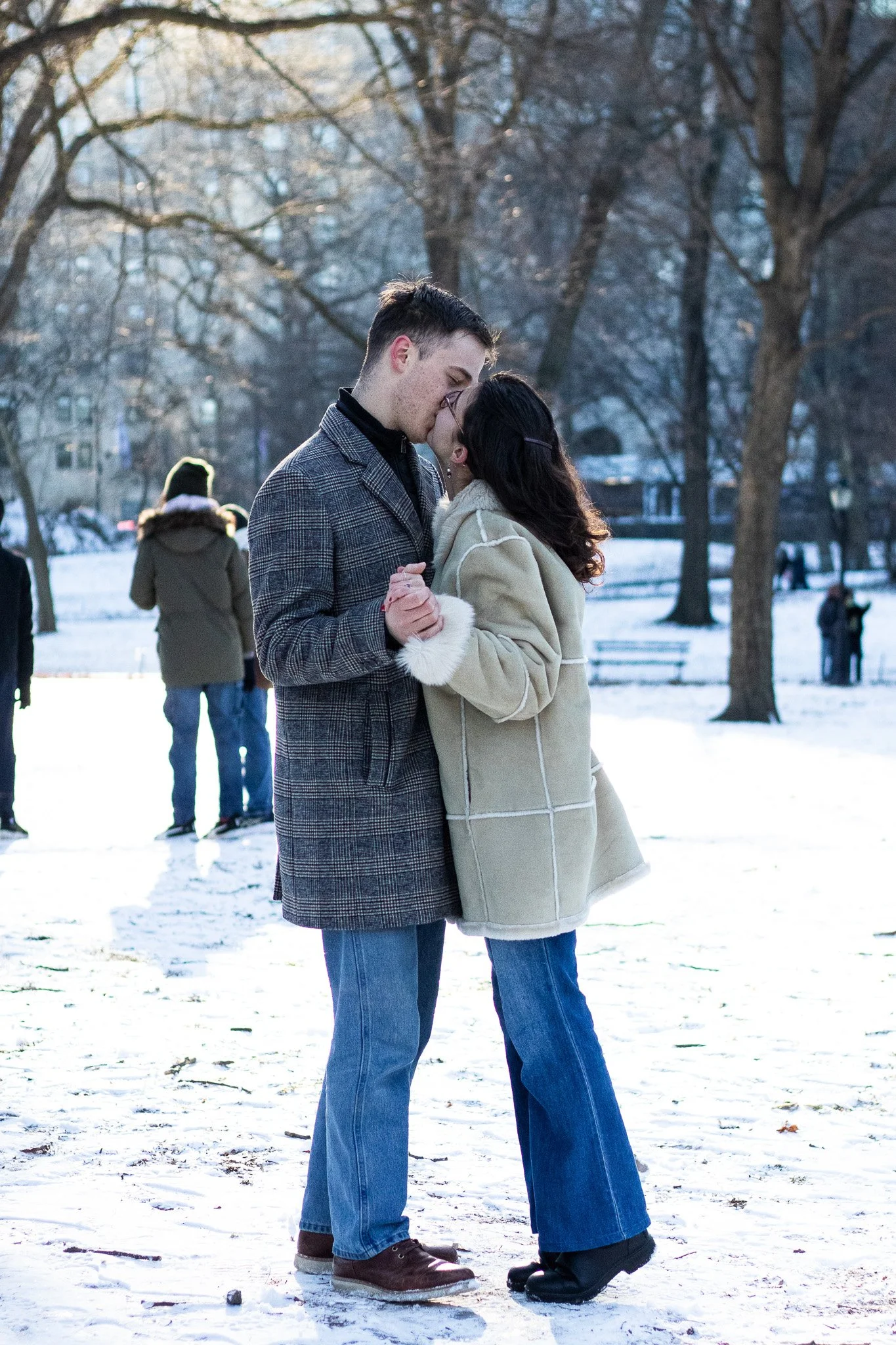 A couple in winter clothing standing close and holding hands in a snowy park, leaning in for a kiss, with other people in the background.