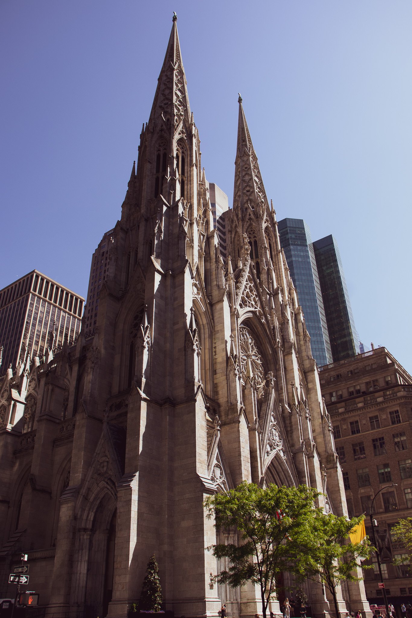Gothic-style church with two tall spires, surrounded by modern skyscrapers, on a sunny day with clear blue sky and some people walking nearby.