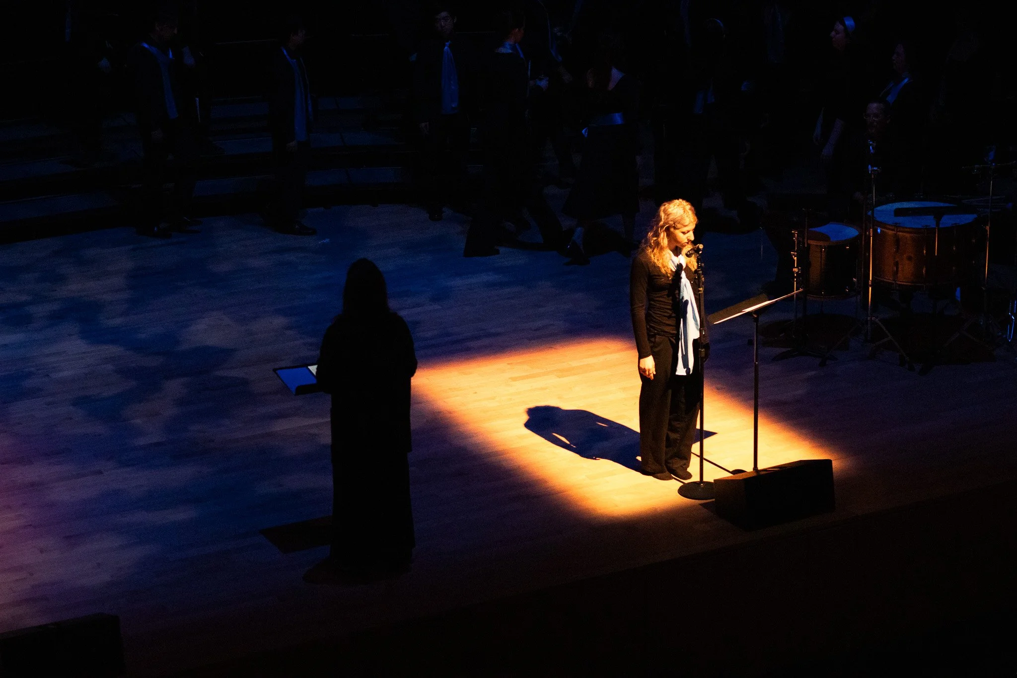 A woman with blonde curly hair standing at a microphone on stage in a spotlight, with a group of people in dark clothing standing behind her in a dimly lit auditorium.