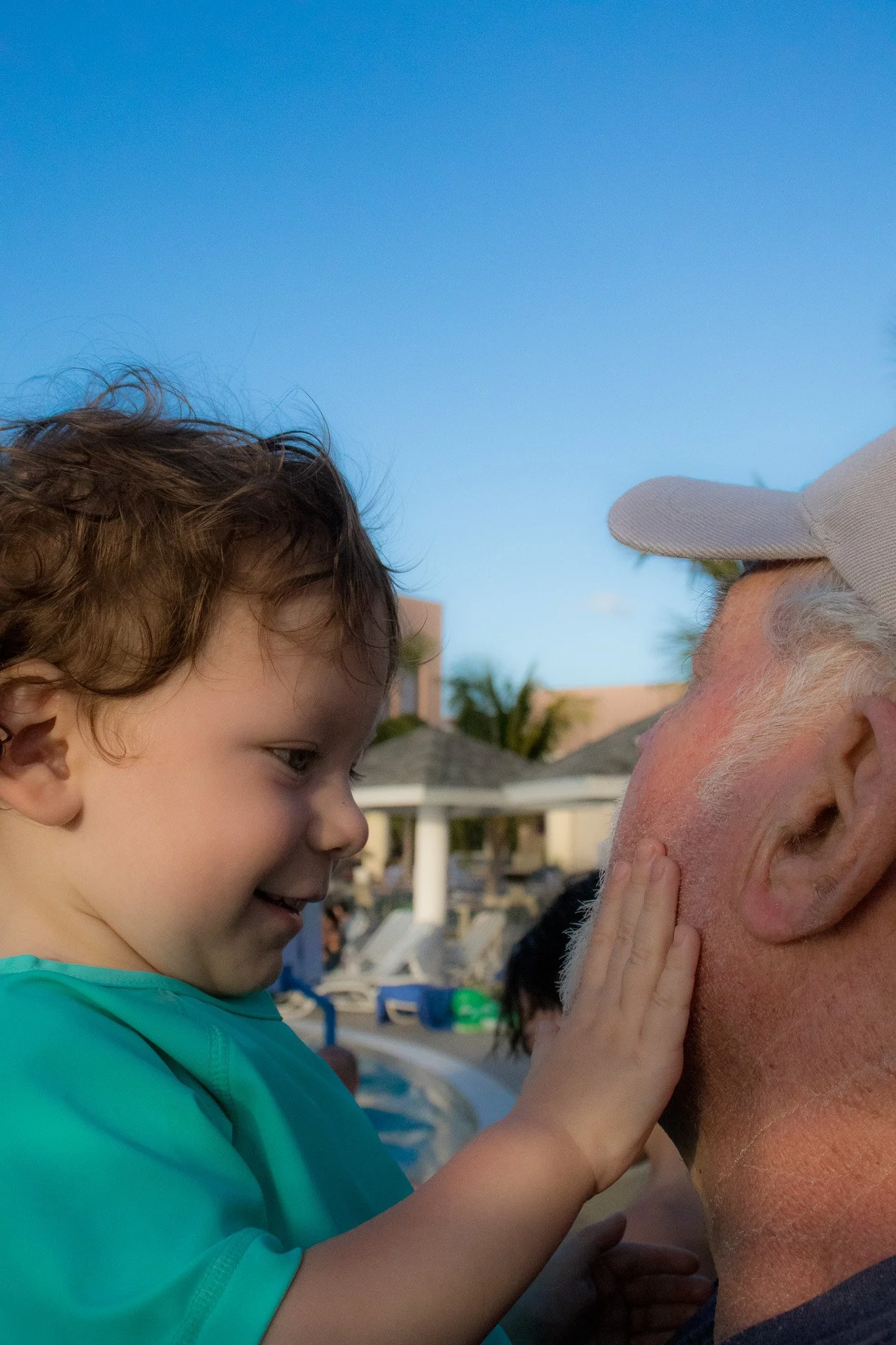 A young child gently touching an older man's face, showing affection in an outdoor setting with blue sky and palm trees in the background.
