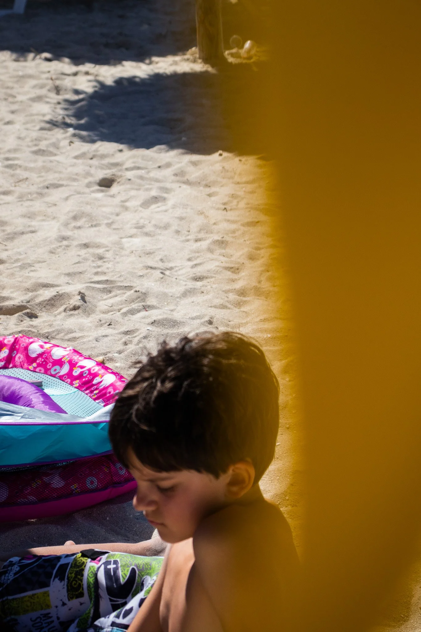 A young boy with brown hair and no shirt sitting on a sandy beach, next to colorful beach toys and a yellow object partially visible on the right side, with shadows and sunlight on the sand.