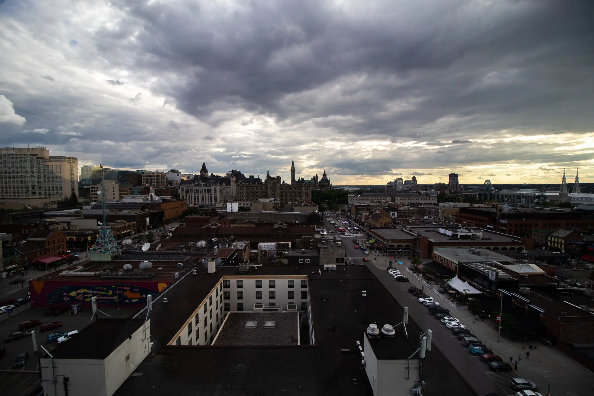 City skyline with tall buildings, some with pointed rooftops, under a cloudy sky with rays of sunlight peeking through.