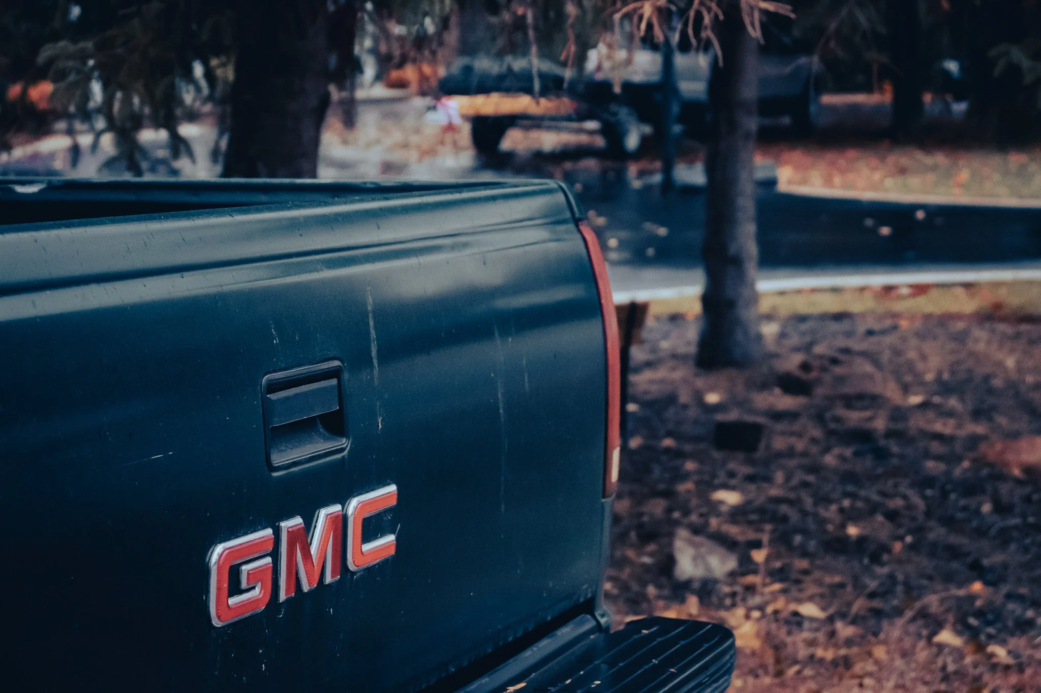 Close-up of the rear of a dark green GMC truck parked outdoors with trees and fallen leaves in the background.