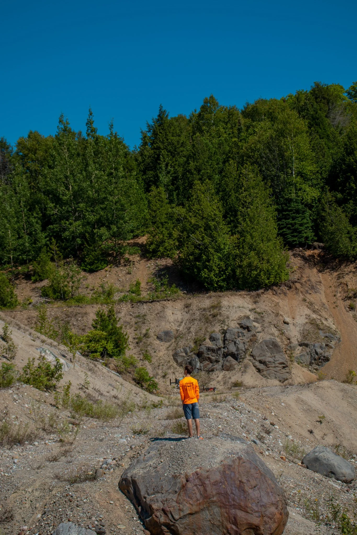 A person in an orange jacket and shorts standing on rocky terrain with a mountain slope, trees, and a clear blue sky in the background.