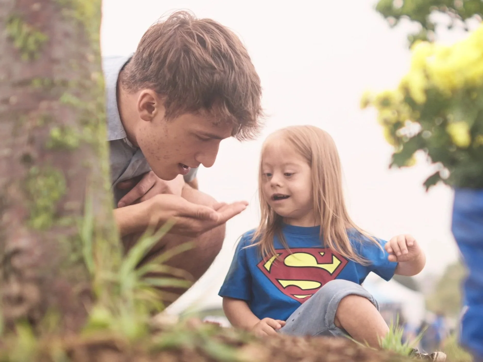 A young man and a girl, dressed as Superman, are outdoors near a tree. The man is showing something on his hand to the girl, and they appear to be curious and happy. The girl has long red hair and is wearing a blue shirt with a Superman logo.
