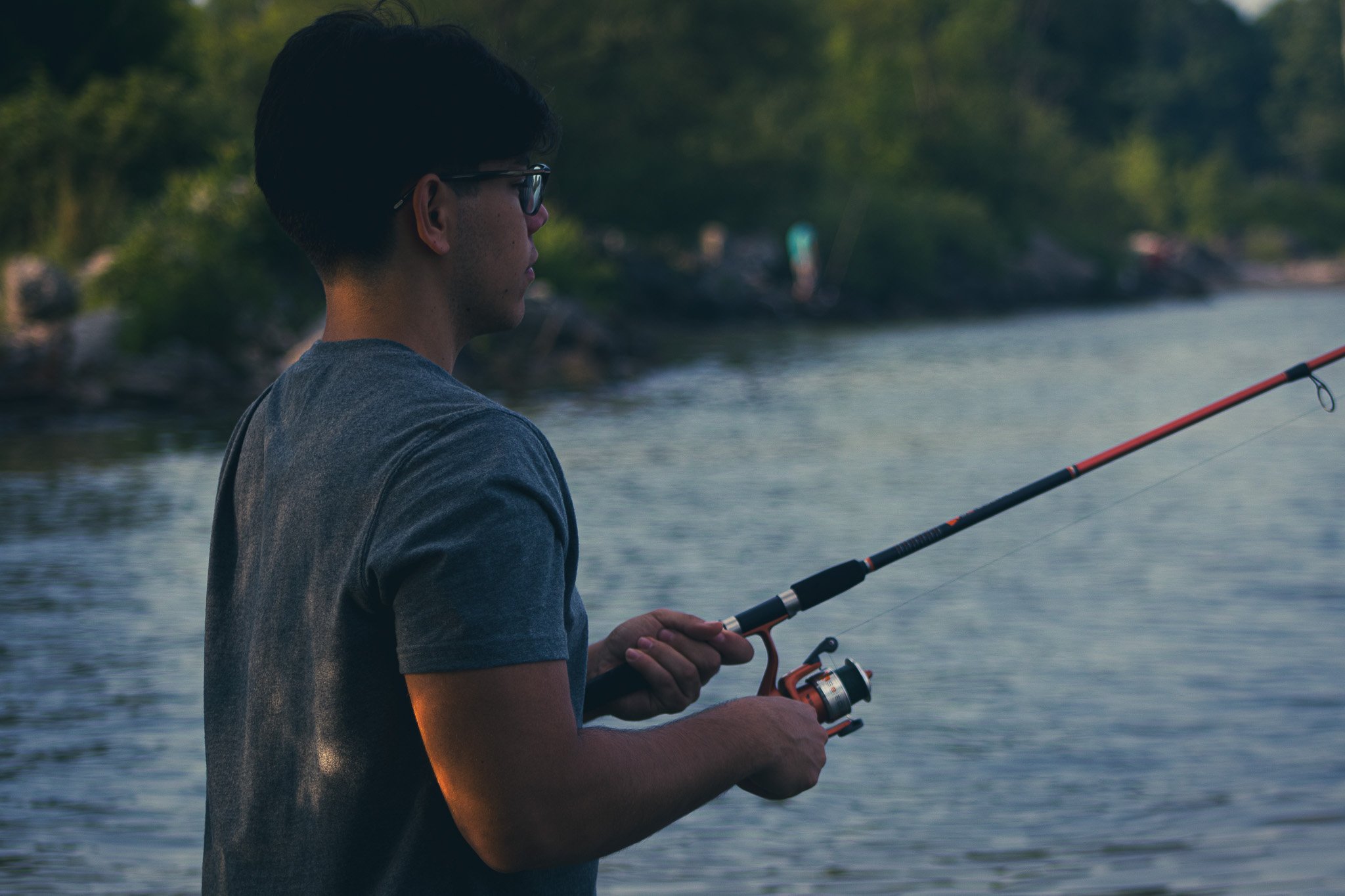 A young man fishing in a lake or river, holding a fishing rod with both hands, wearing sunglasses and a gray t-shirt, with trees and water in the background.
