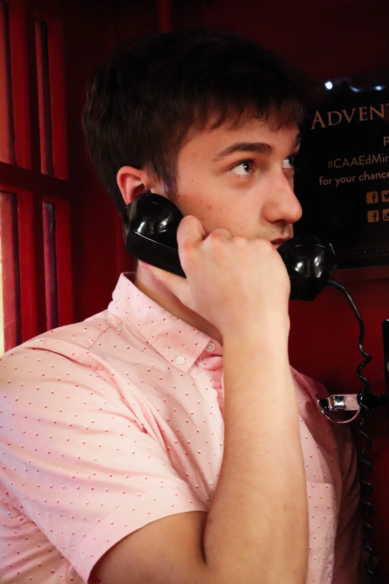 A young man with dark hair and light skin holding a black telephone receiver to his ear, wearing a pink shirt with small red dots, in front of a red wooden background and a black sign with partially visible text.