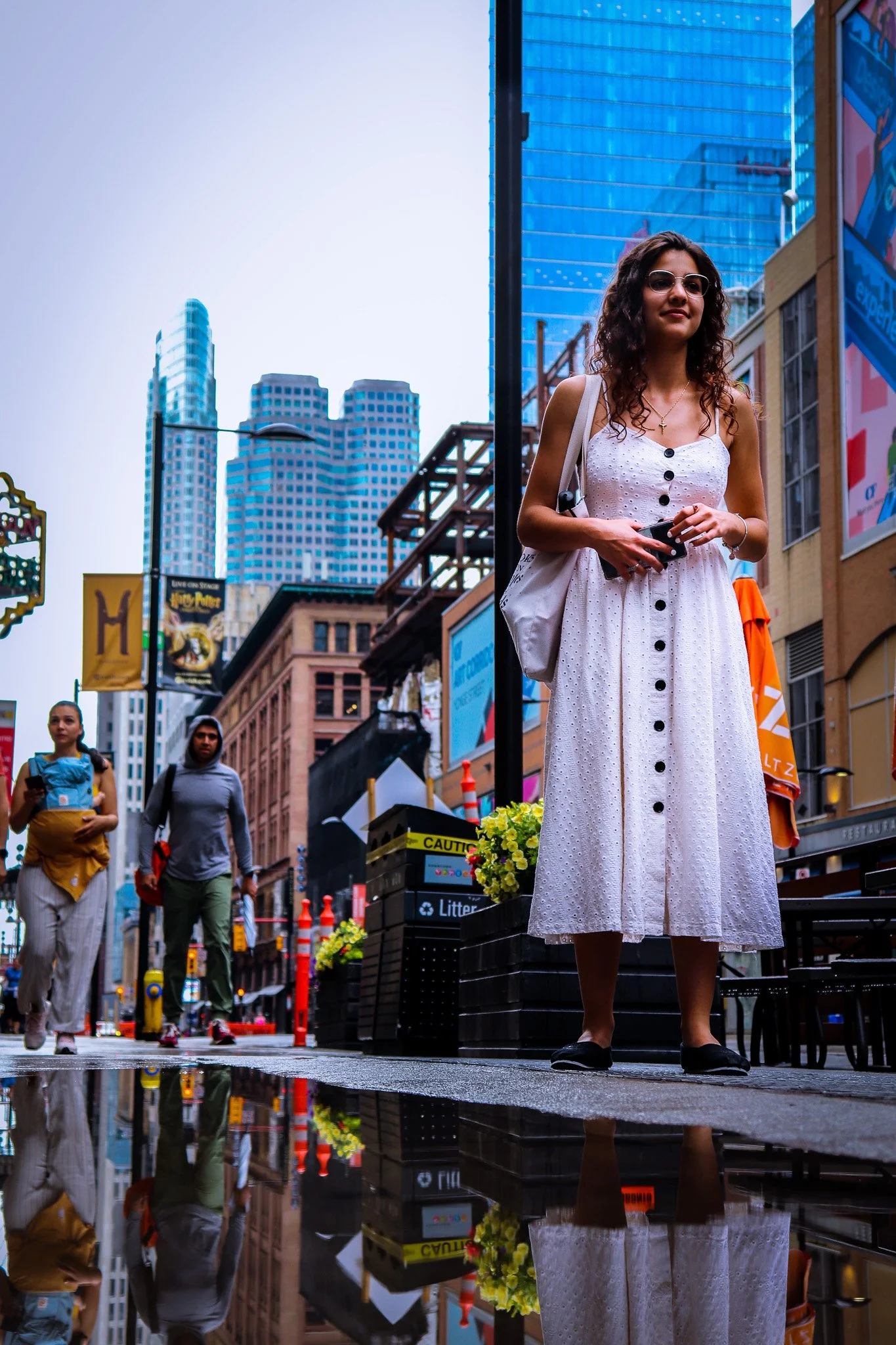 A woman wearing sunglasses and a white dress with black buttons standing on a city street with skyscrapers, other pedestrians, and reflections on wet pavement.