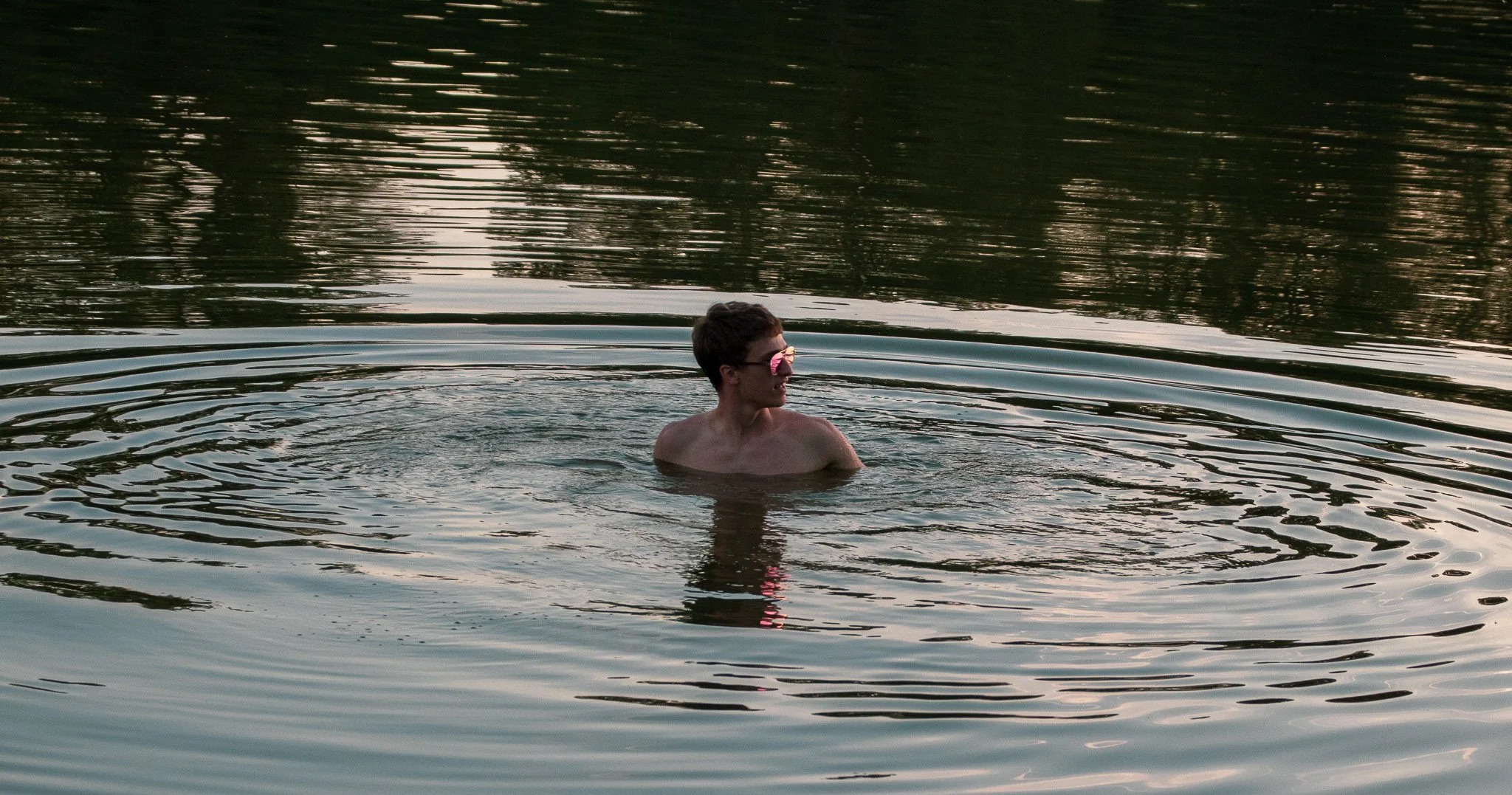 A young man with dark hair and sunglasses swimming in a body of water during sunset or sunrise, with reflecting ripples on the water surface.
