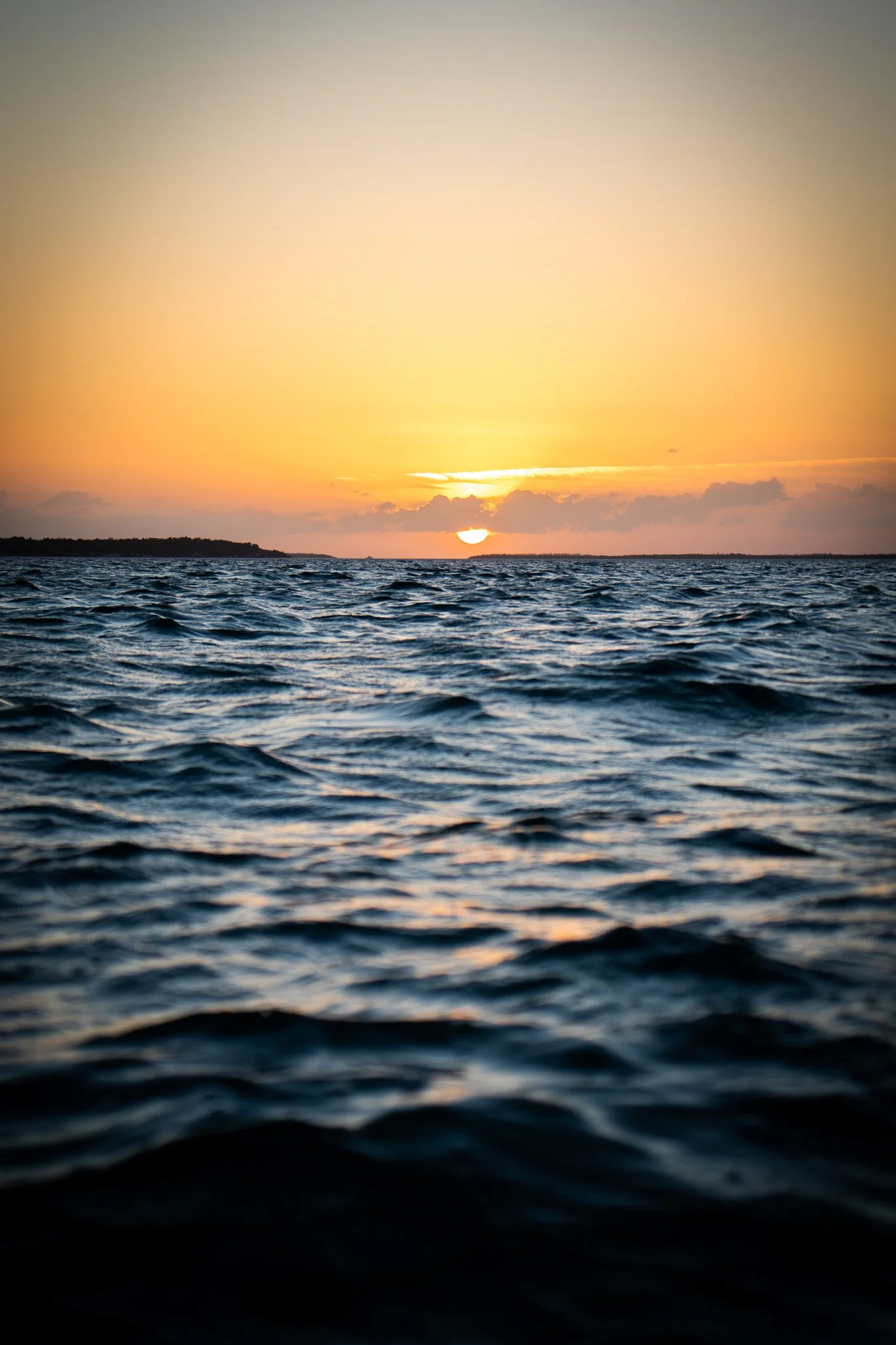 Sunset over the ocean with a distant shoreline, featuring a calm and rippled water surface and a partly cloudy sky.