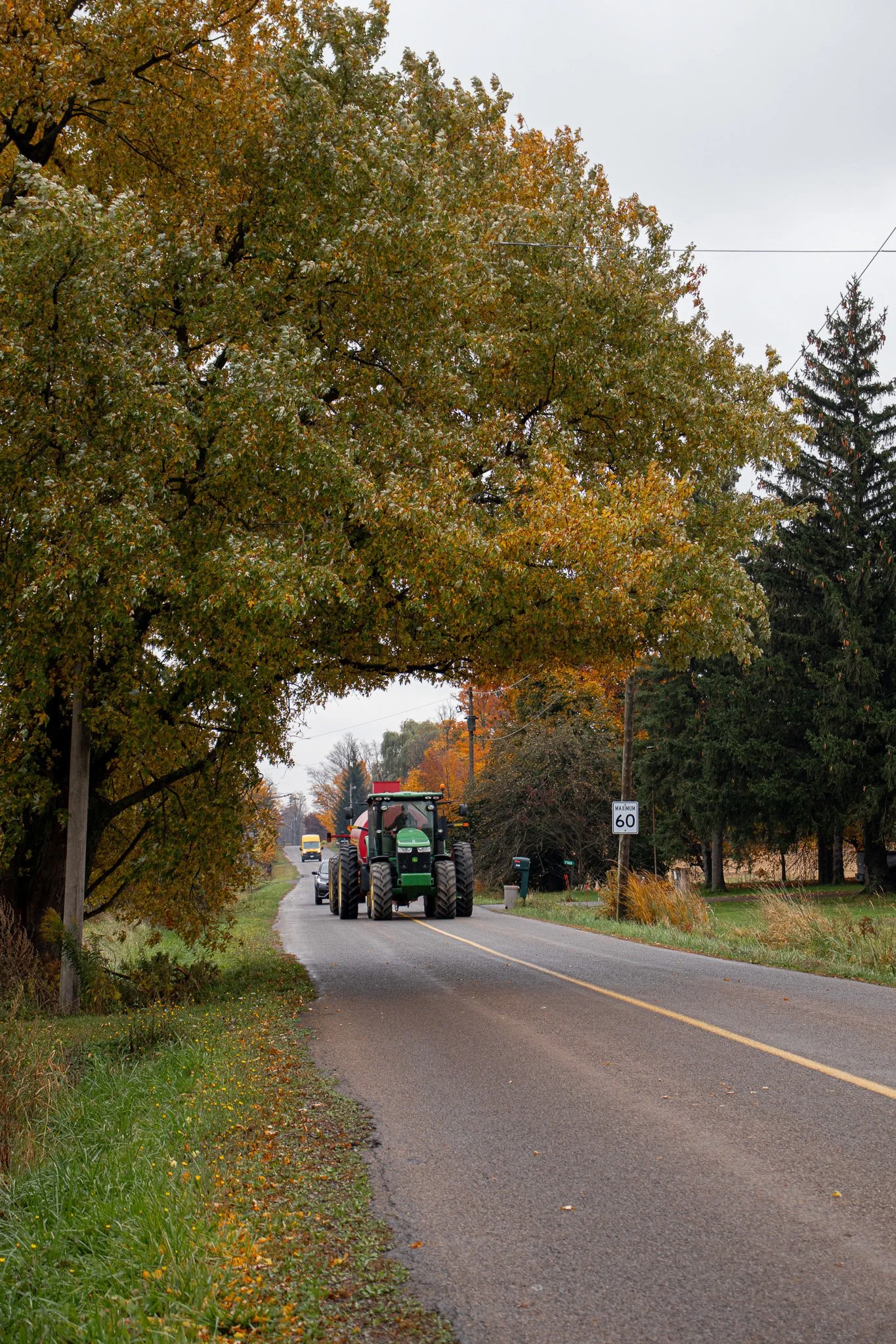 A rural street scene during fall with a large green tractor driving on a narrow paved road, surrounded by colorful trees and a gray overcast sky.