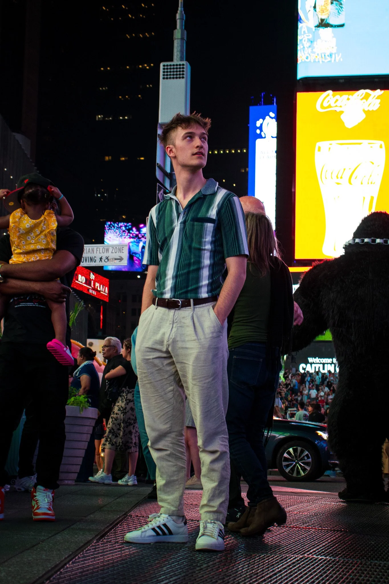 A young man stands in Times Square at night, with neon billboards and digital screens illuminating the background. He wears a striped shirt, light-colored trousers, and striped sneakers, and has a serious expression. There are other people around, in