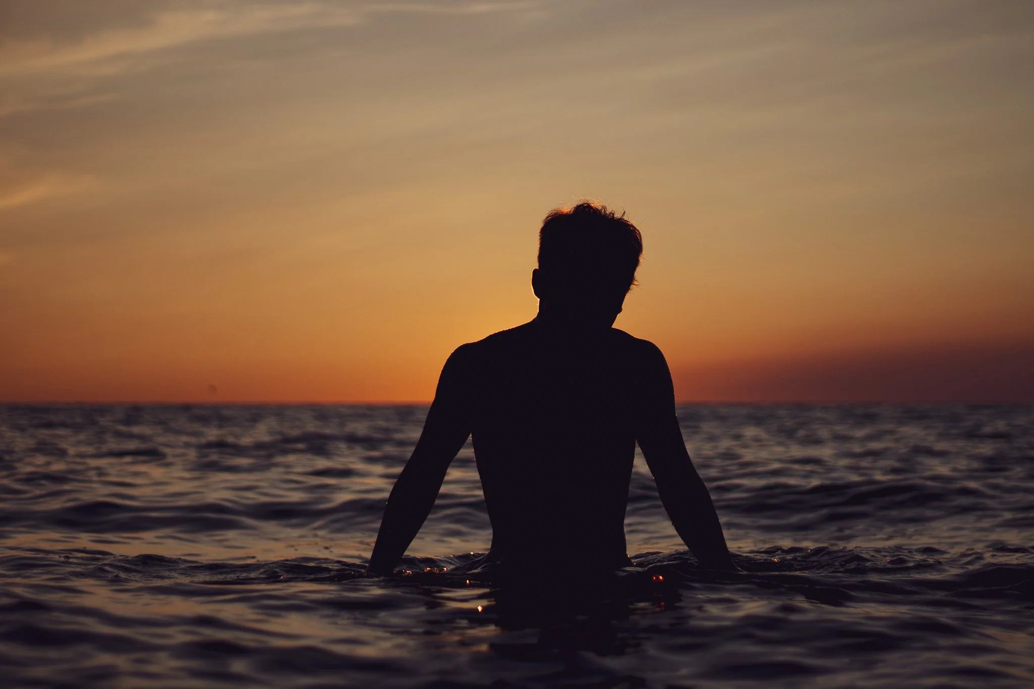 Silhouette of a person sitting in the ocean at sunset, facing the horizon with a sky painted in warm colors and calm water.