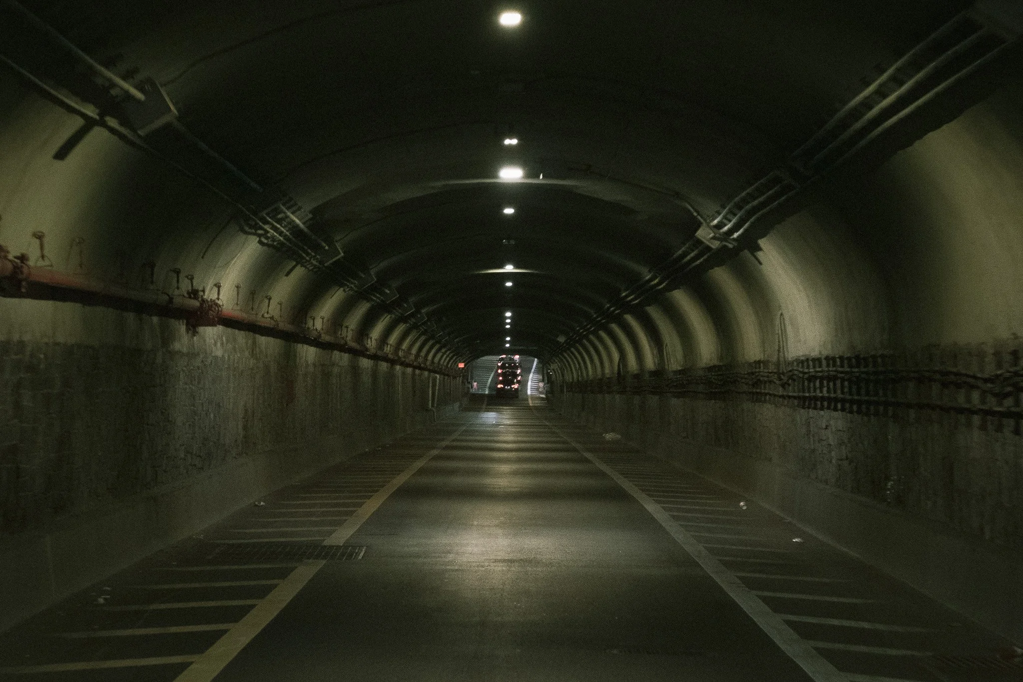 Underwater tunnel with curved ceiling, illuminated by ceiling lights, with a vehicle entering or exiting at the far end.