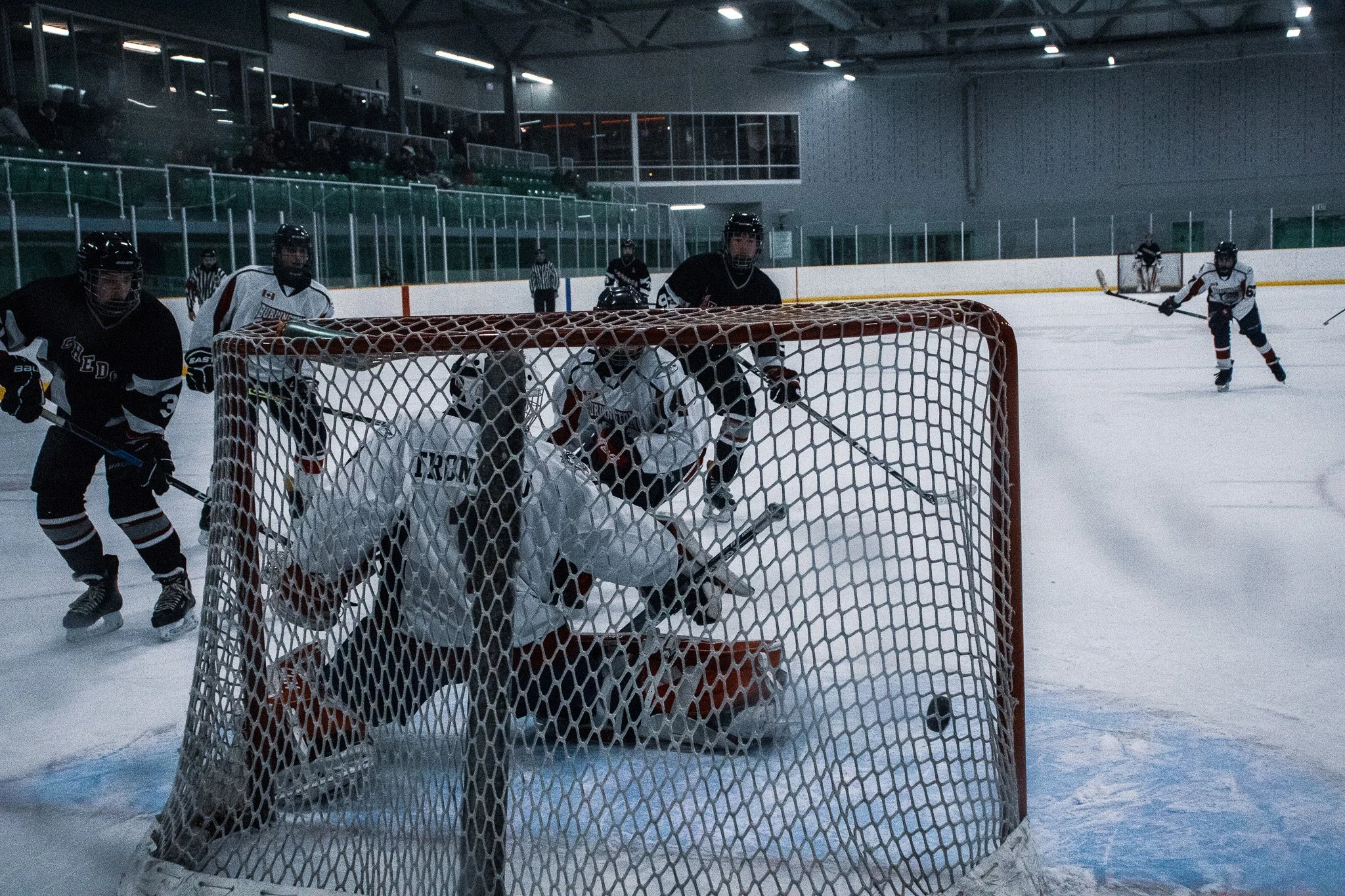 Ice hockey game in progress with players near the goal, some wearing black and some white jerseys, inside an indoor ice rink with spectators in the stands.