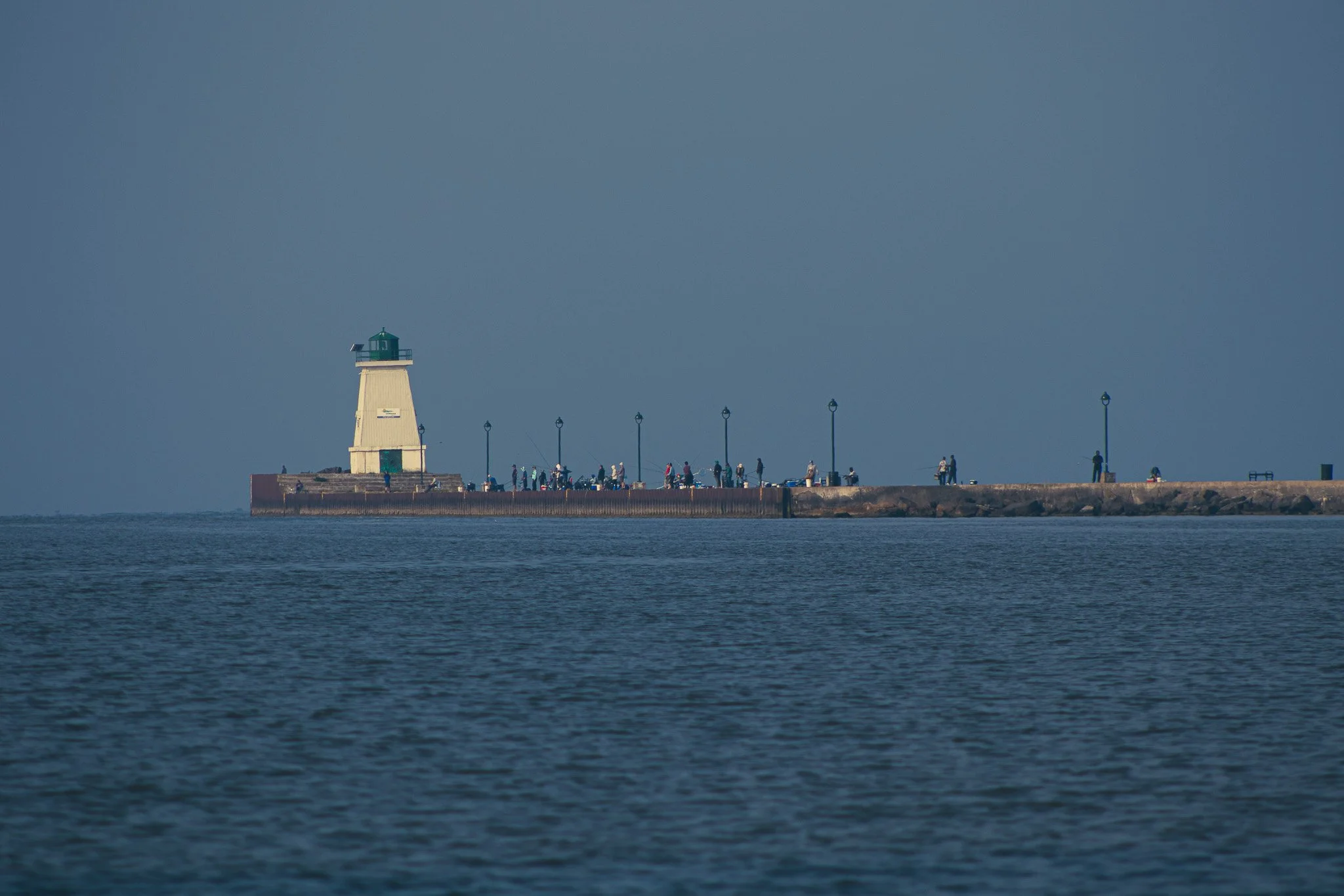 A lighthouse on a pier with people fishing and walking, against a blue sky and ocean.