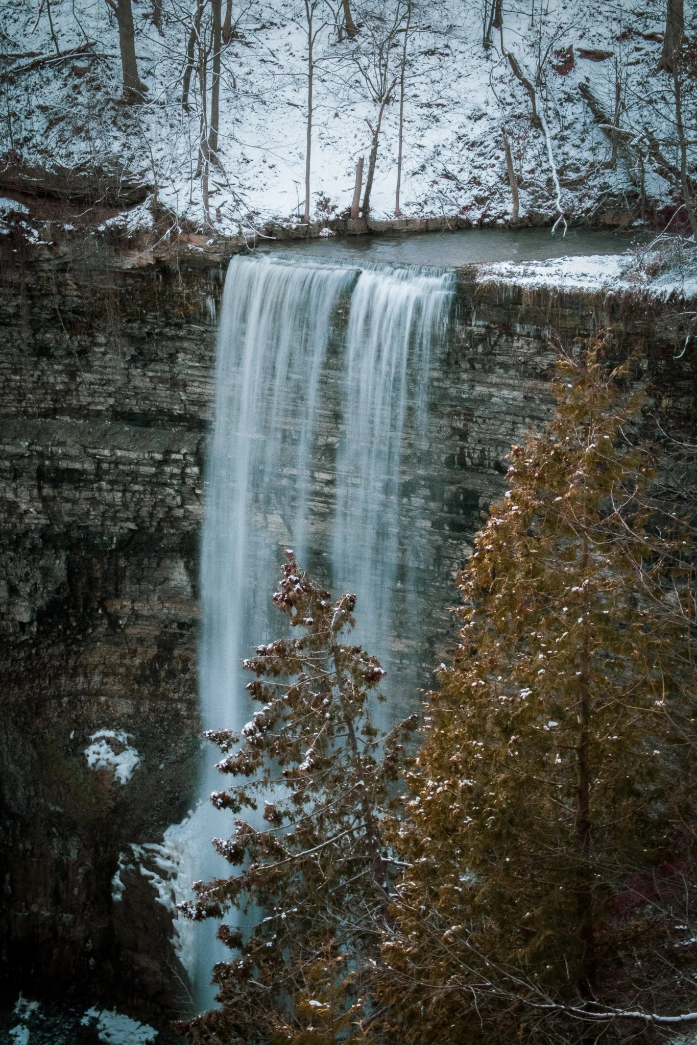 Waterfall flowing over rocks in a wintery forest with snow-covered trees and branches.