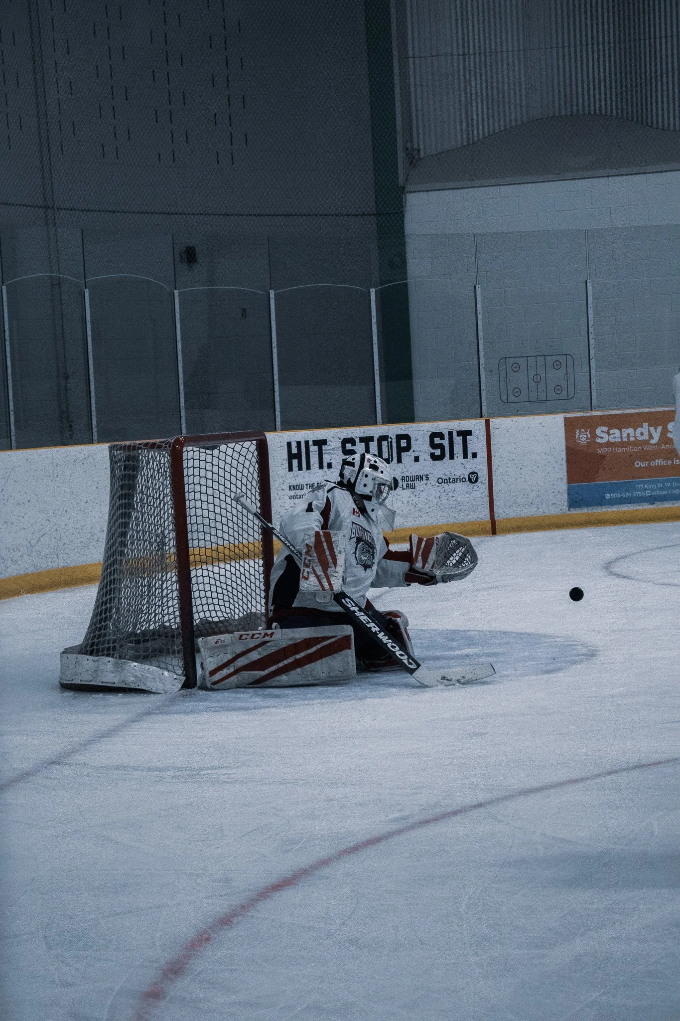 Hockey goalie in white gear sitting in front of the net on ice rink, preparing to block a puck.