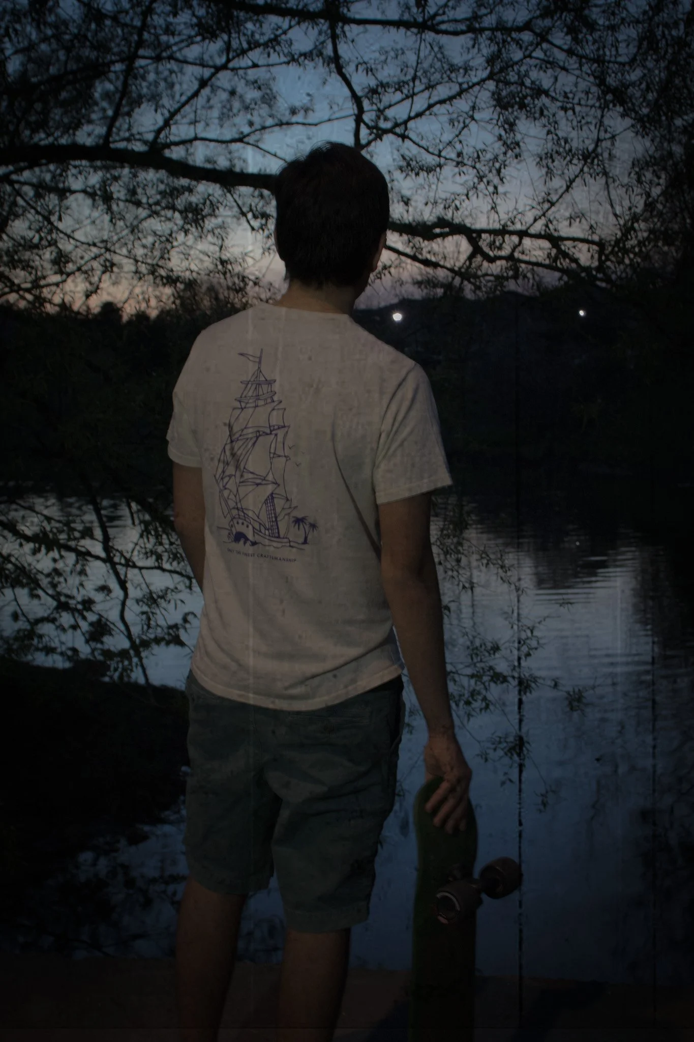 A person standing near a body of water at dusk or dawn, holding a skateboard in one hand, with trees and mountain in the background.