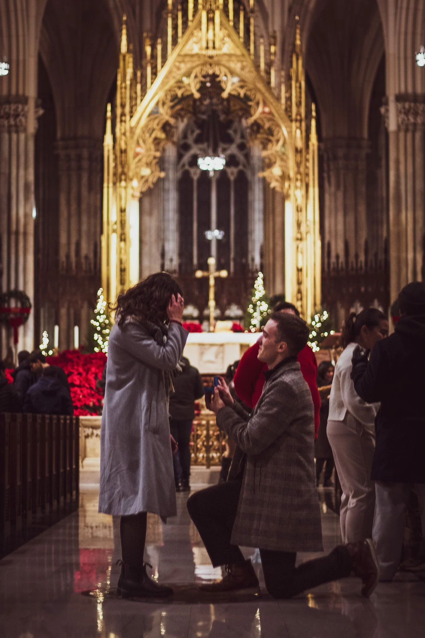A man kneeling and proposing to a woman inside a church decorated for Christmas, with people around watching.