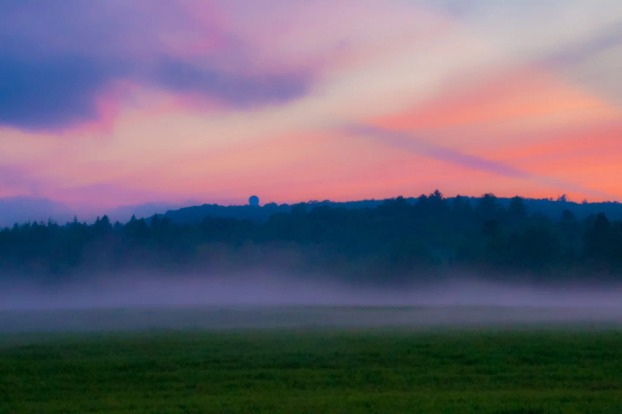 A landscape view of a sunset or sunrise with a colorful sky in shades of pink, purple, and blue, and a silhouette of trees and a water tower on the horizon.