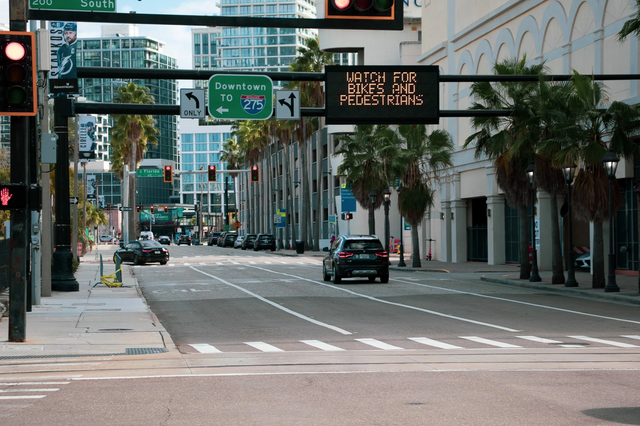 City street with palm trees, traffic lights, and a digital signboard displaying 'Watch for bikes and pedestrians.' Tall buildings are visible in the background, including a banner with a hockey player and the text 'CKA.' A green road sign indicates d