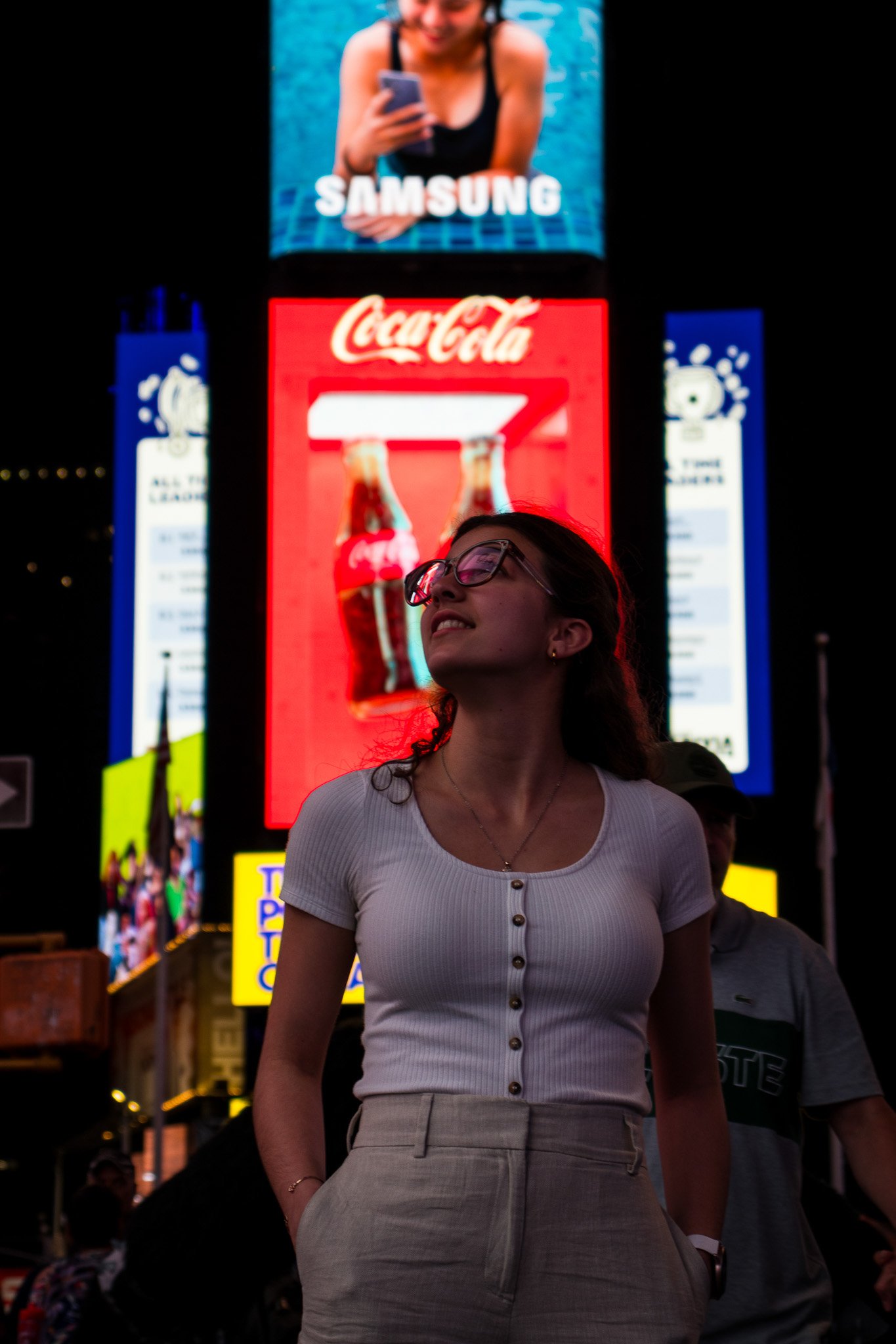 A woman with curly hair and glasses stands in Times Square at night, illuminated by bright advertisements, including a Samsung ad at the top and a Coca-Cola ad below.