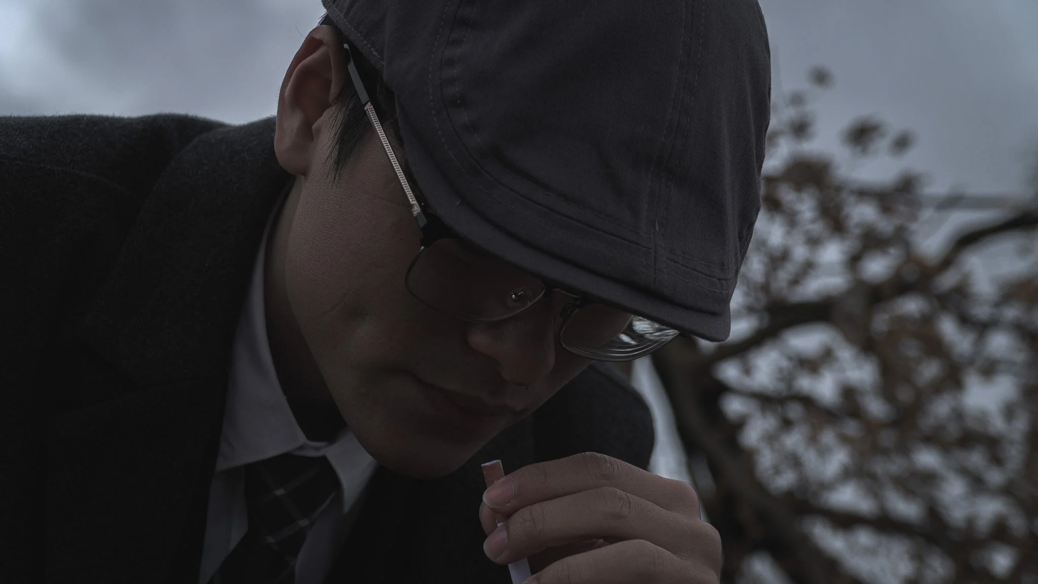 A person wearing a black cap, glasses, and a dark coat, holding a small object in their hand, with a background of trees and a cloudy sky.