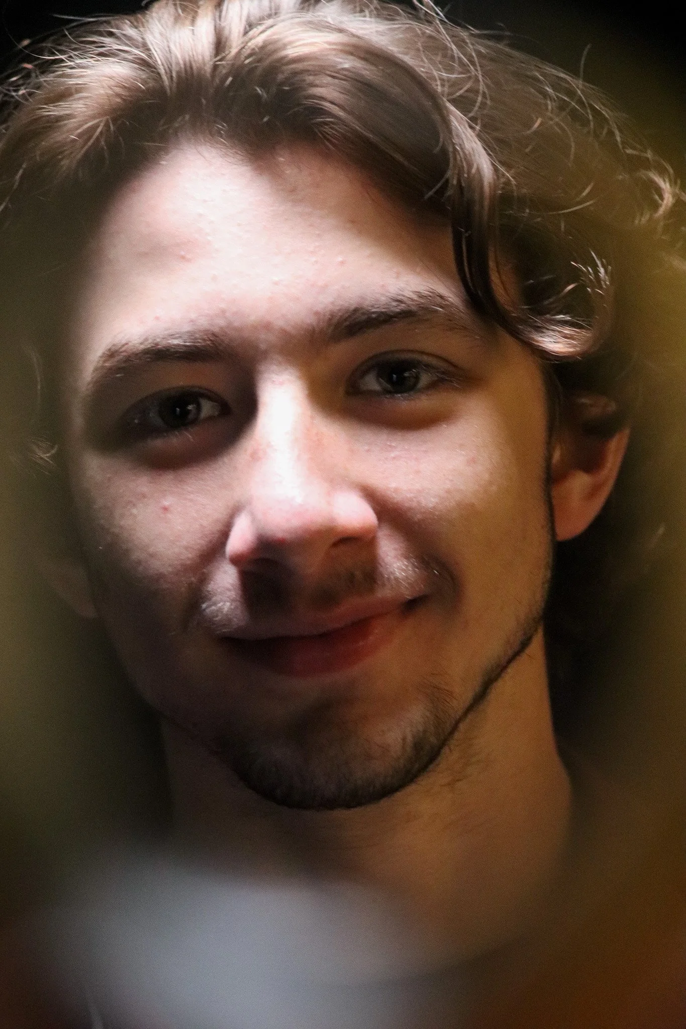 Close-up of a young man's face with dark hair, brown eyes, and a faint smile, partially obscured by a blurred foreground.