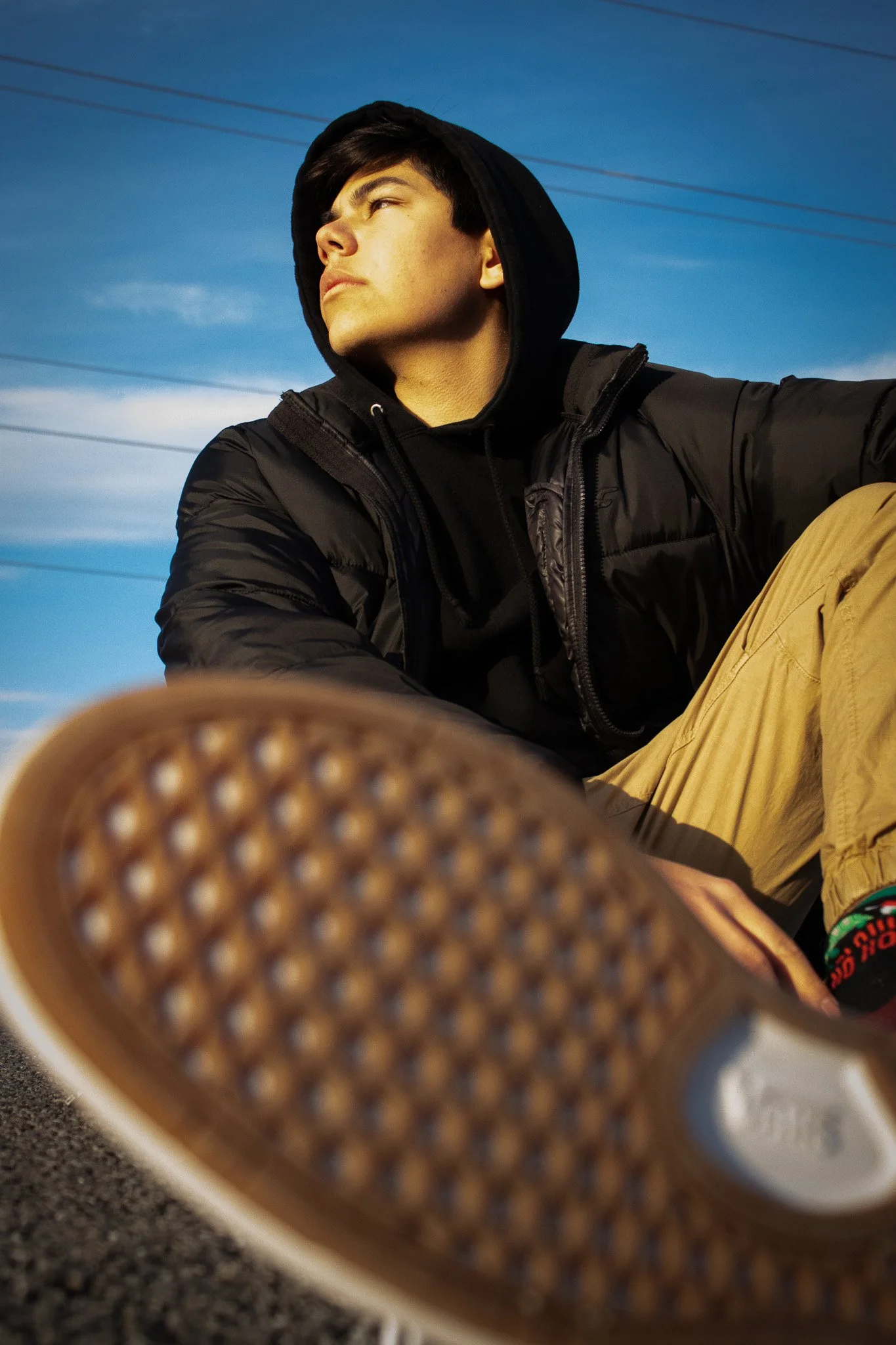 A young man with dark hair wearing a black hoodie and tan pants sitting outdoors, with power lines in the background, and foot in the foreground wearing a sneaker with a patterned sole.