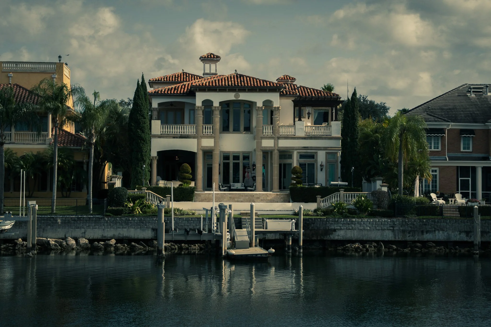 Luxury waterfront house with a tiled roof, lush greenery, and a dock extending over calm water.