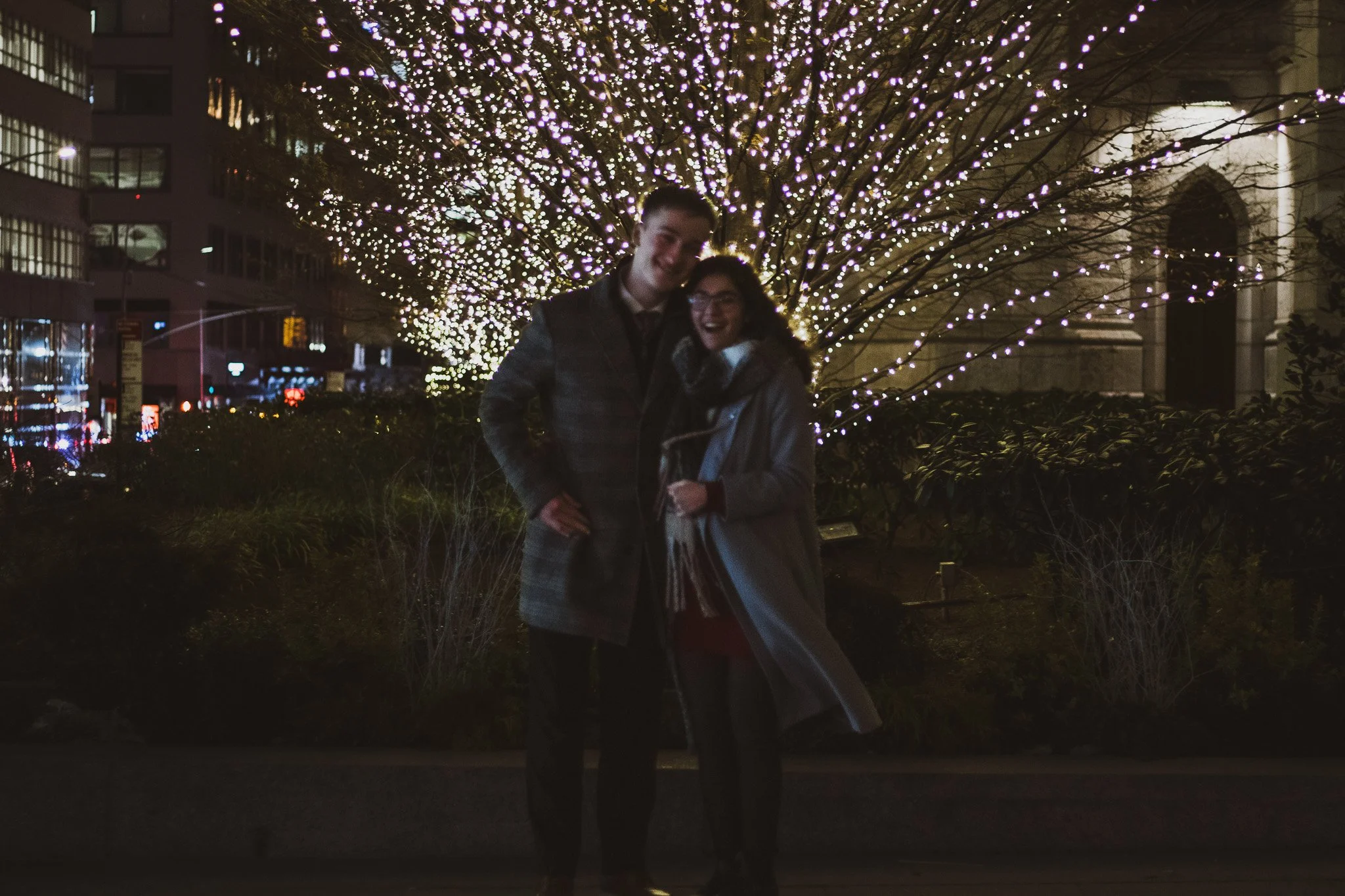 Night scene with two smiling people, a man and a woman, standing in front of a large tree decorated with purple and white string lights. City buildings and streetlights are visible in the background.