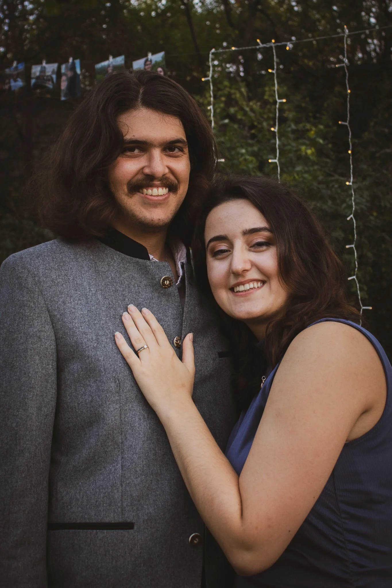 A smiling man with long dark hair, a mustache, and a gray coat, standing outdoors with a woman who has wavy dark hair and is wearing a sleeveless navy dress. They are close together, smiling at the camera, with string lights and photographs hanging i