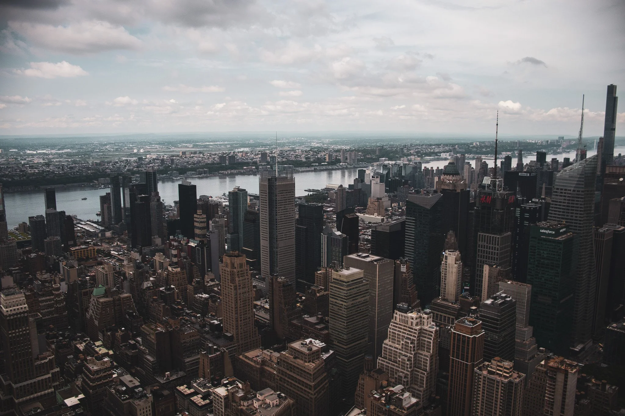 A panoramic view of a city skyline featuring numerous tall skyscrapers, a wide river, numerous buildings, and a cloudy sky.