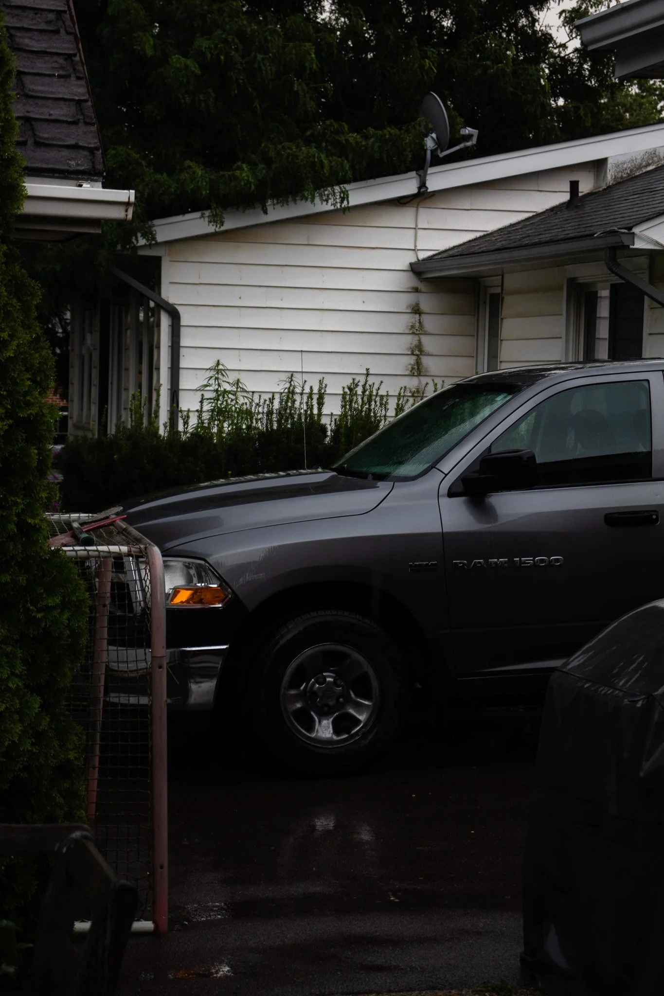 A gray pickup truck parked outside a residential house with white siding and dark roofs. The house has a satellite dish on the roof, and there are some plants and trees around.