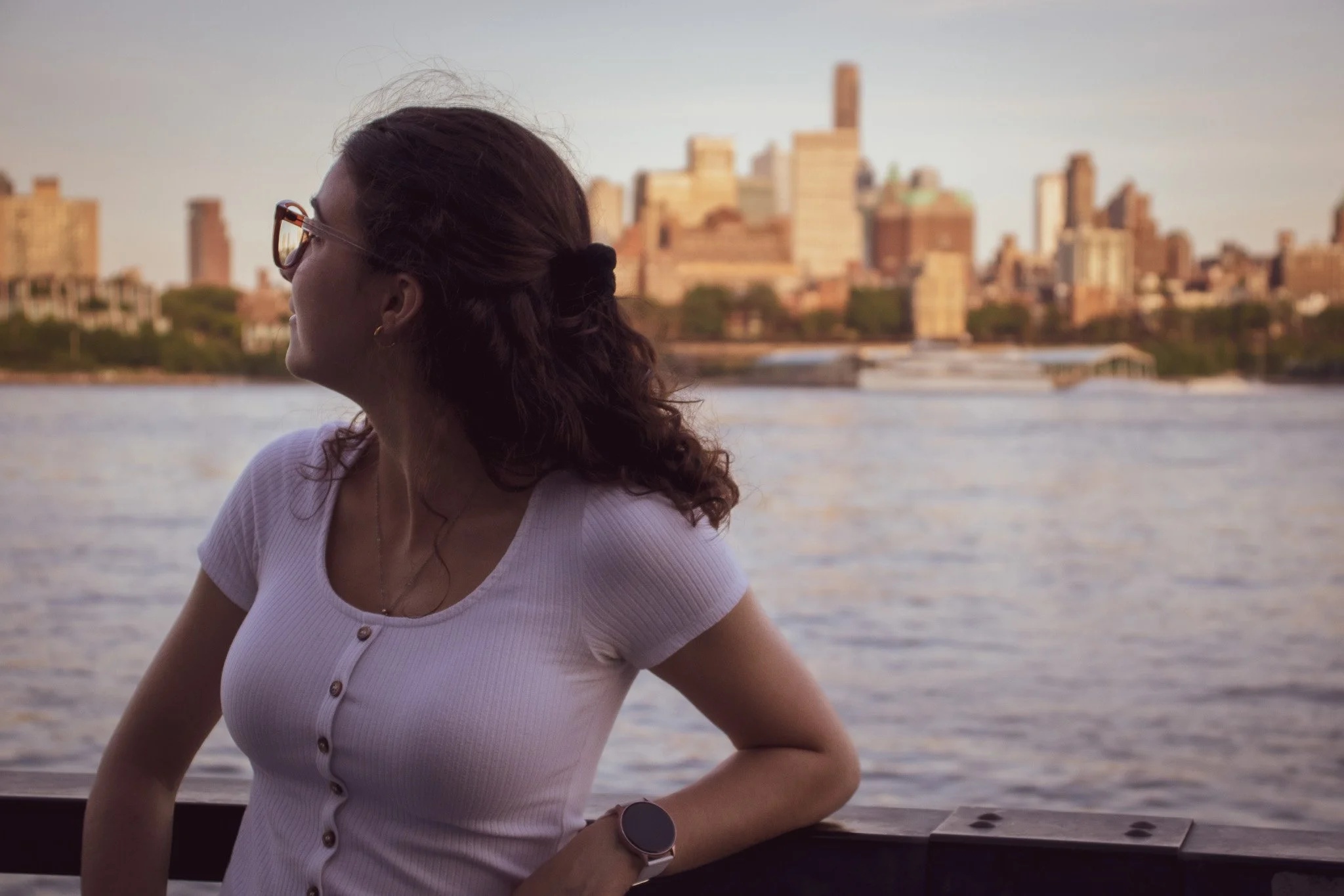 A woman with dark wavy hair tied back with a black hair tie, wearing glasses, a white button-up shirt, and a smartwatch, is looking to the left with a city skyline in the background. The scene appears to be near a body of water during sunset.