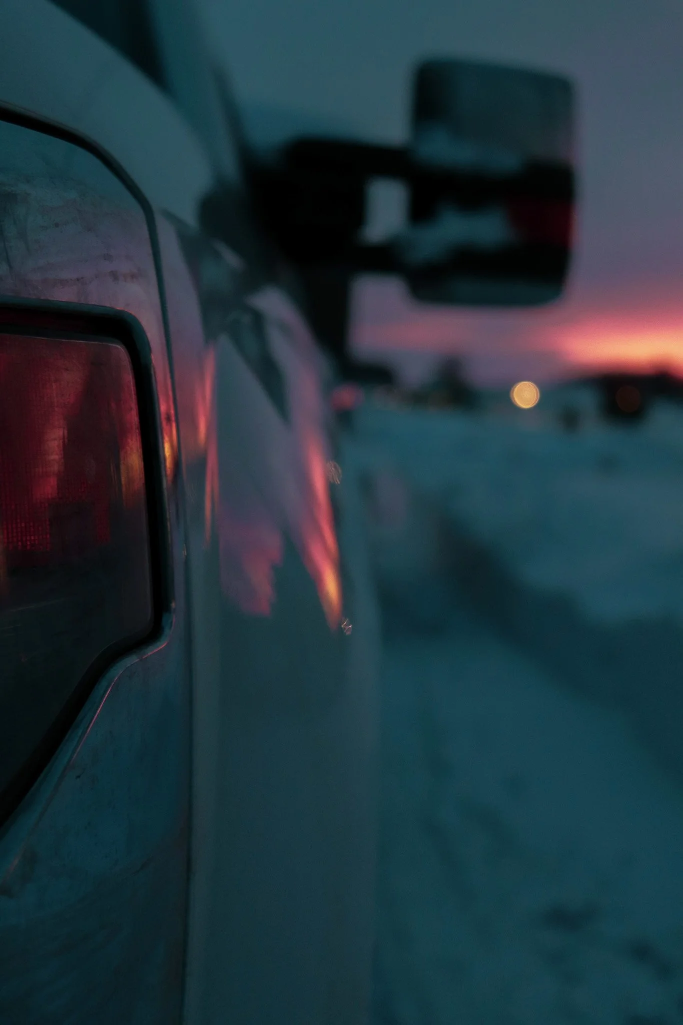 Close-up of a car's rear with a sunset in the background, focusing on the car's tail light and side mirror.