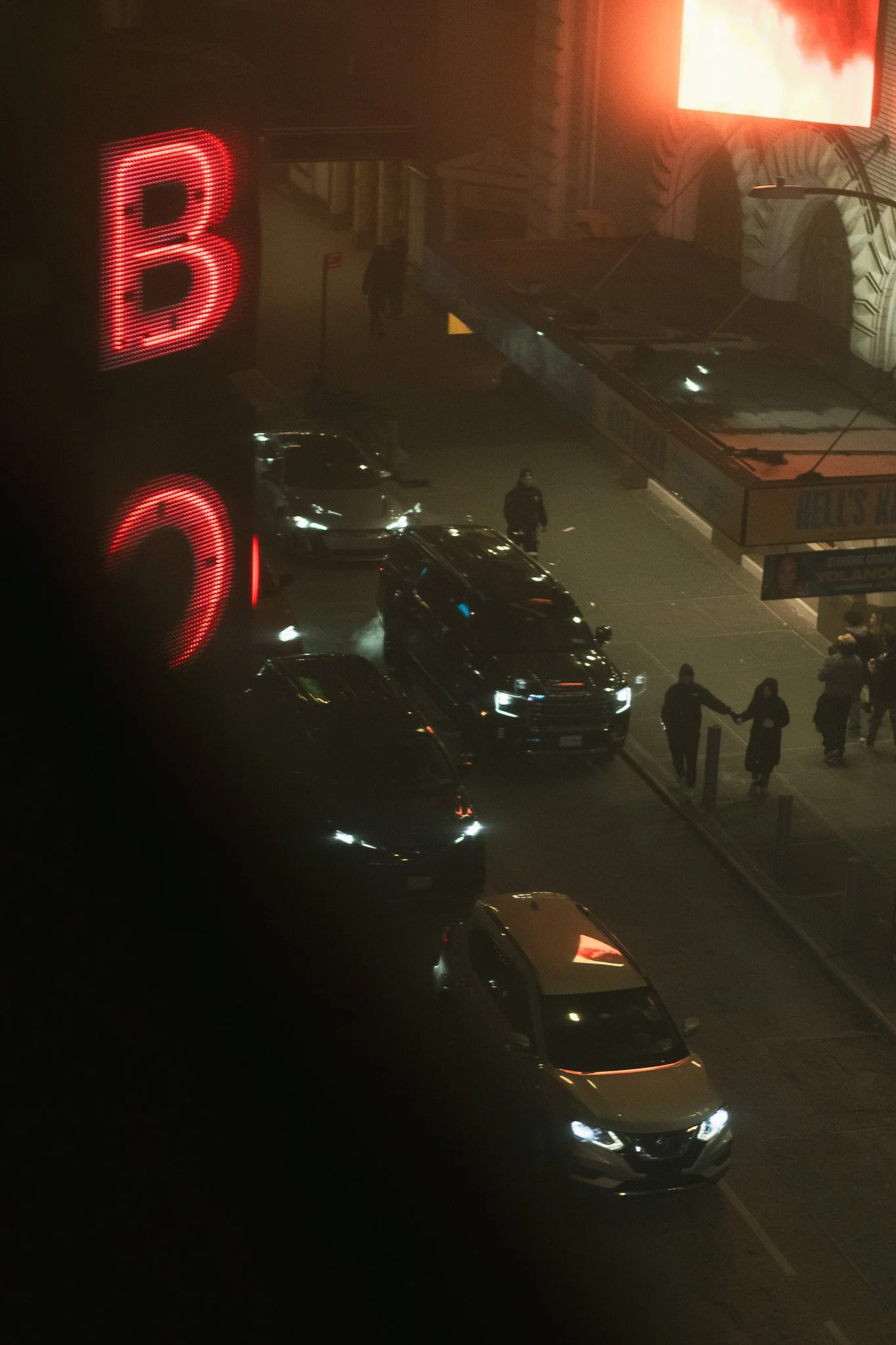 Nighttime city scene with bright red neon letter 'B', several cars with headlights on, and a person walking on the sidewalk. There is a large illuminated billboard in the background and a street lamp.