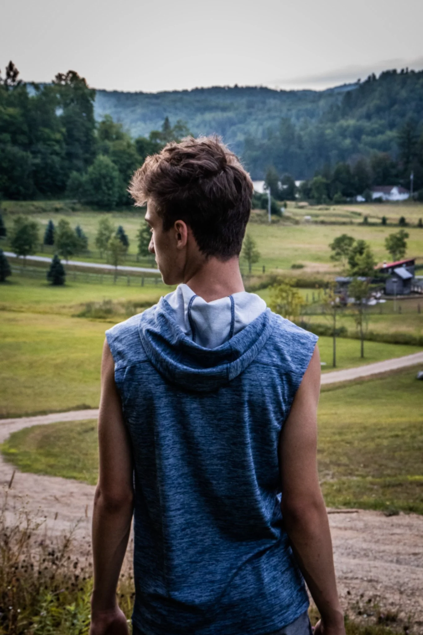 Young man with short brown hair wearing a sleeveless blue hoodie, standing outdoors on a dirt path, looking towards a scenic landscape of rolling green hills, trees, and mountains in the distance during daytime.
