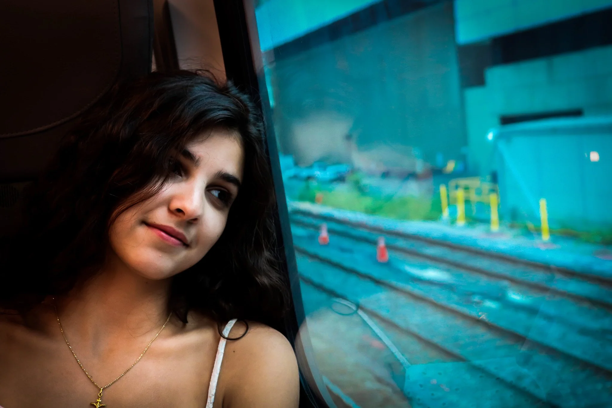 A young woman with long, dark curly hair looking out a bus window, with a slight smile on her face. She is wearing a white spaghetti strap top and a gold necklace with a star pendant. The bus window shows a blurred view of the street outside.