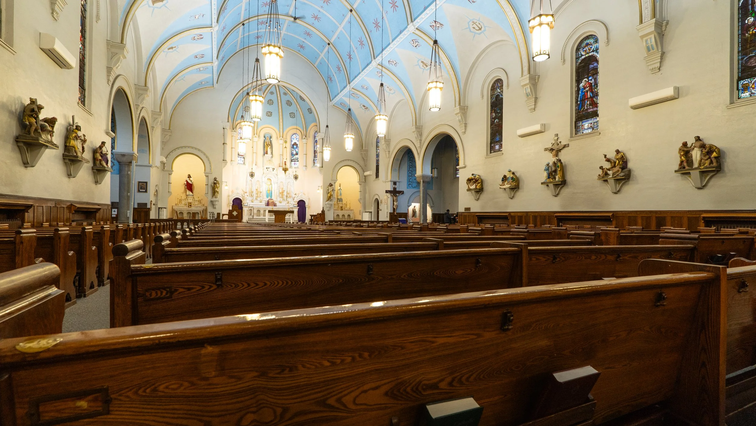 Interior of a church with wooden pews, a high arched ceiling painted light blue with stars and symbols, stained glass windows, and religious statues and artwork on the walls.