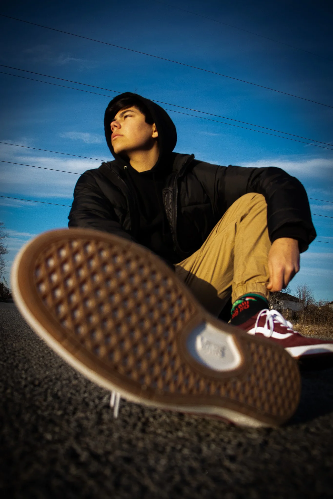 A young man in a black hoodie and tan pants sitting on a skateboard on the street, with a low angle perspective focusing on his sneaker, with a blue sky and power lines in the background.
