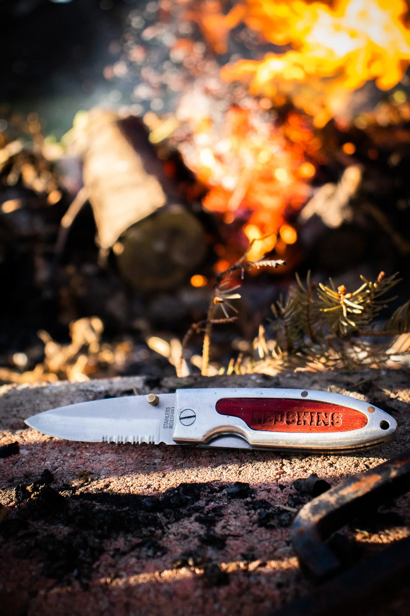A close-up photo of a folding pocket knife with a partially serrated blade lying on the ground, with a fire burning in the background.